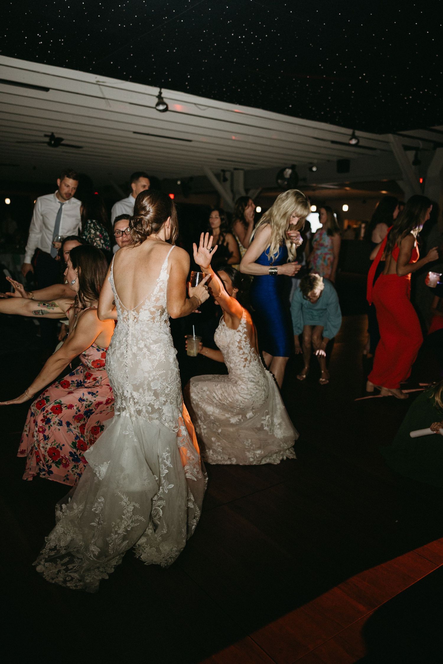 Two brides in lace gowns dance with guests at lively wedding reception.film-inspired