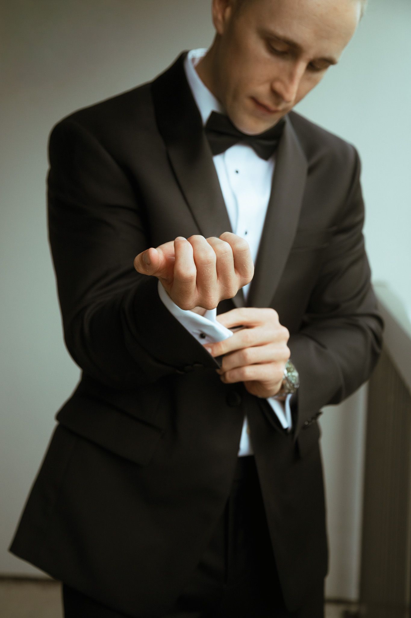 Groom in black tuxedo with bow tie fastening cufflinks while getting ready for wedding.