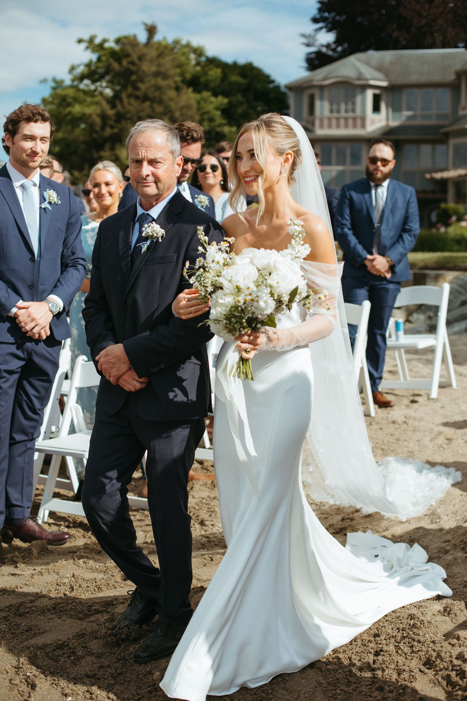 Bride walking down sandy beach aisle with father, holding white floral bouquet during outdoor ceremony.