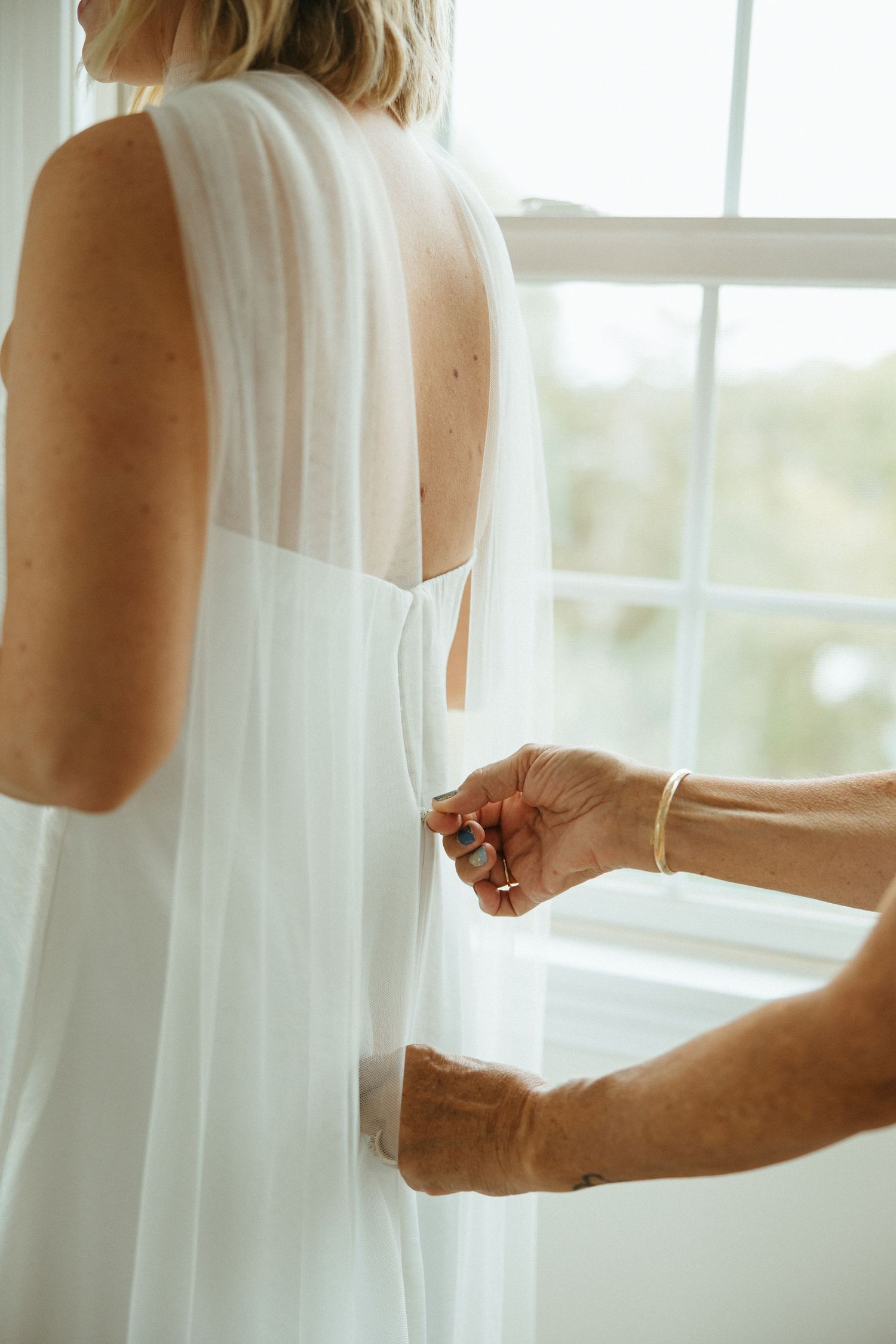 Helper fastening buttons on back of white wedding dress near bright window.