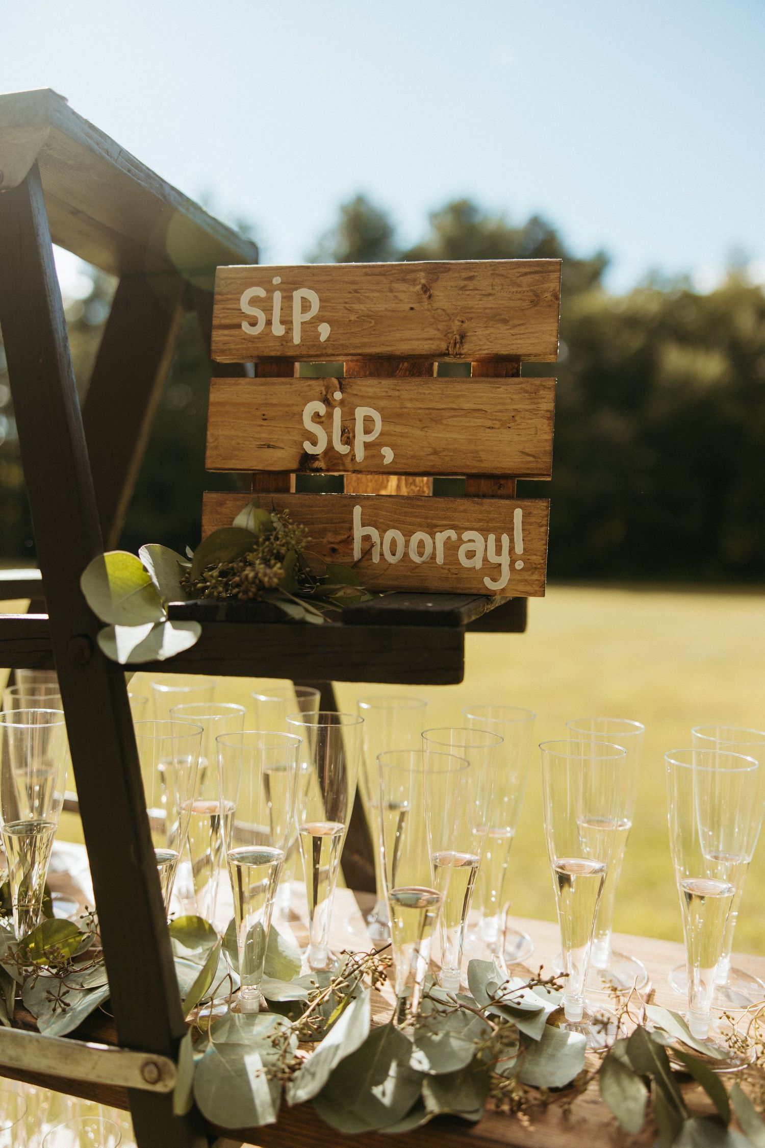 Rustic wooden sip sip hooray sign displayed above champagne flutes at an outdoor wedding reception.