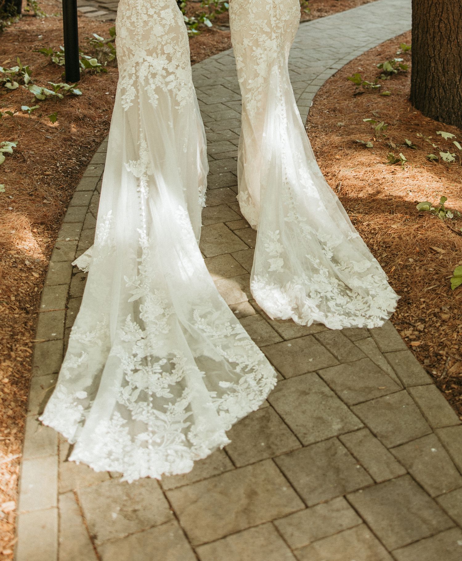 Two brides in matching lace gowns with long trains walk along brick pathway after ceremony.
