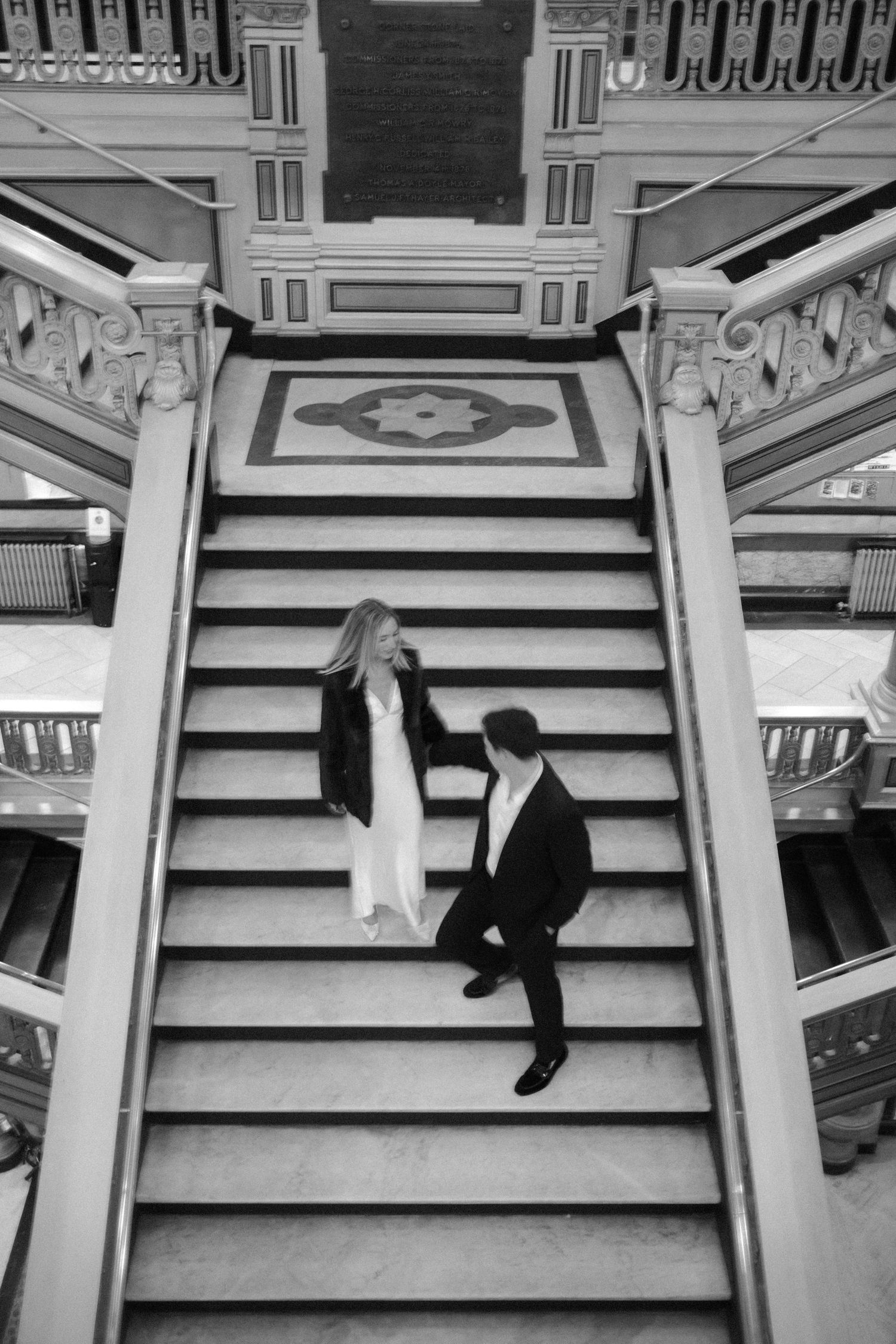 Aerial black and white photo of a couple sitting together on grand ornate staircase in historic building.film-inspired