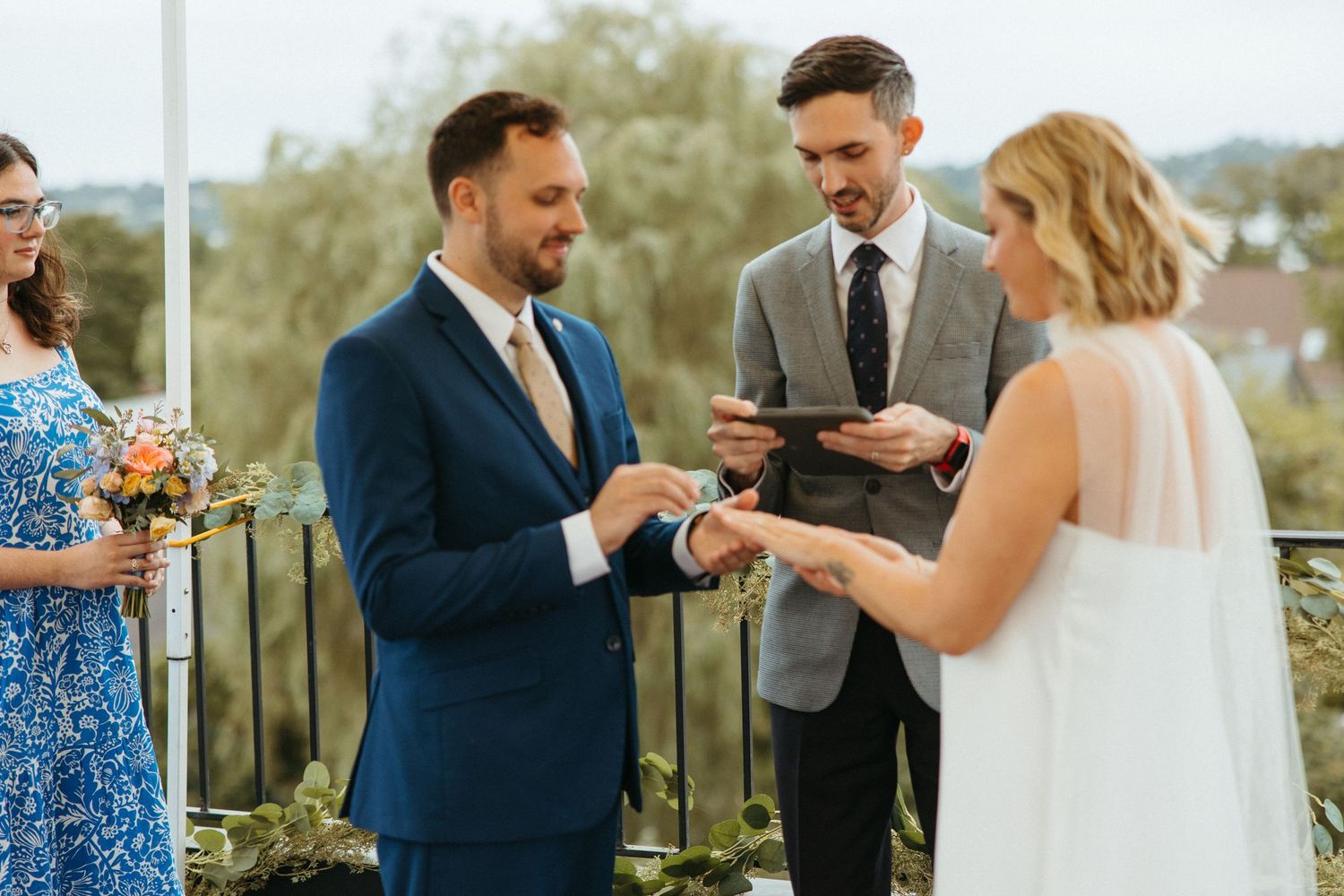 Groom places wedding ring on bride's finger during outdoor rooftop ceremony with officiant and bridesmaid.