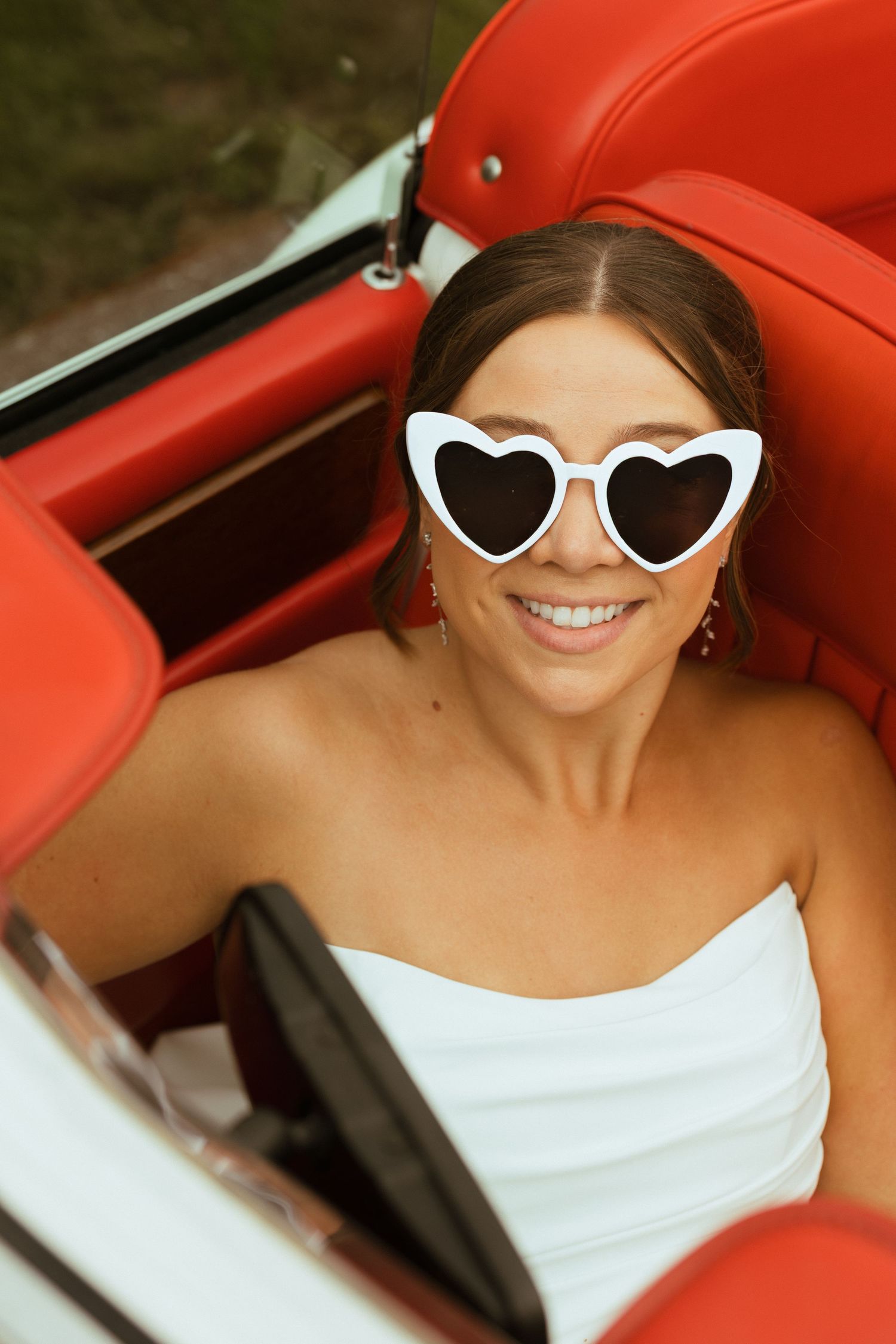 Smiling bride wearing white heart-shaped sunglasses sits in red interior of a classic convertible car.