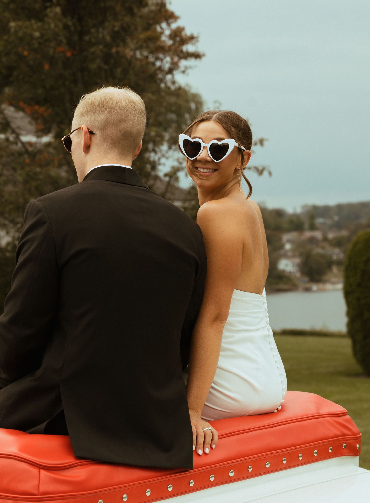 Bride wearing heart-shaped sunglasses smiling while seated on a vintage red and white convertible car.