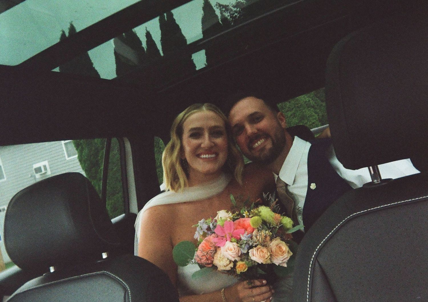 Smiling bride and groom sitting together in vehicle with floral bouquet on wedding day.
