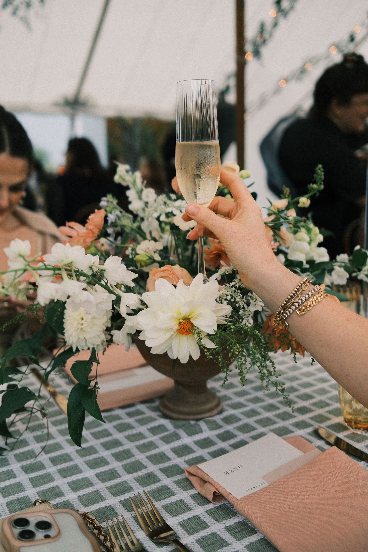Hand holding a champagne flute near a white and peach floral centerpiece at an elegant tented wedding reception.
