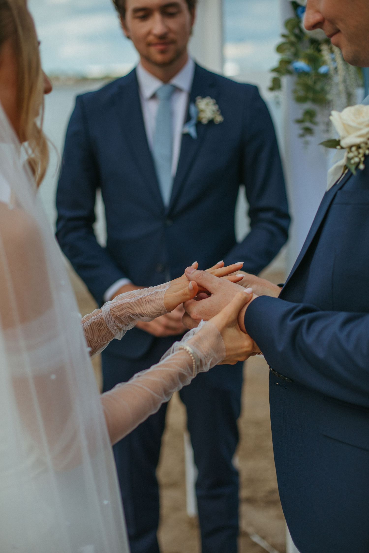 Close-up of bride placing wedding ring on groom's finger during outdoor beach ceremony with officiant.
