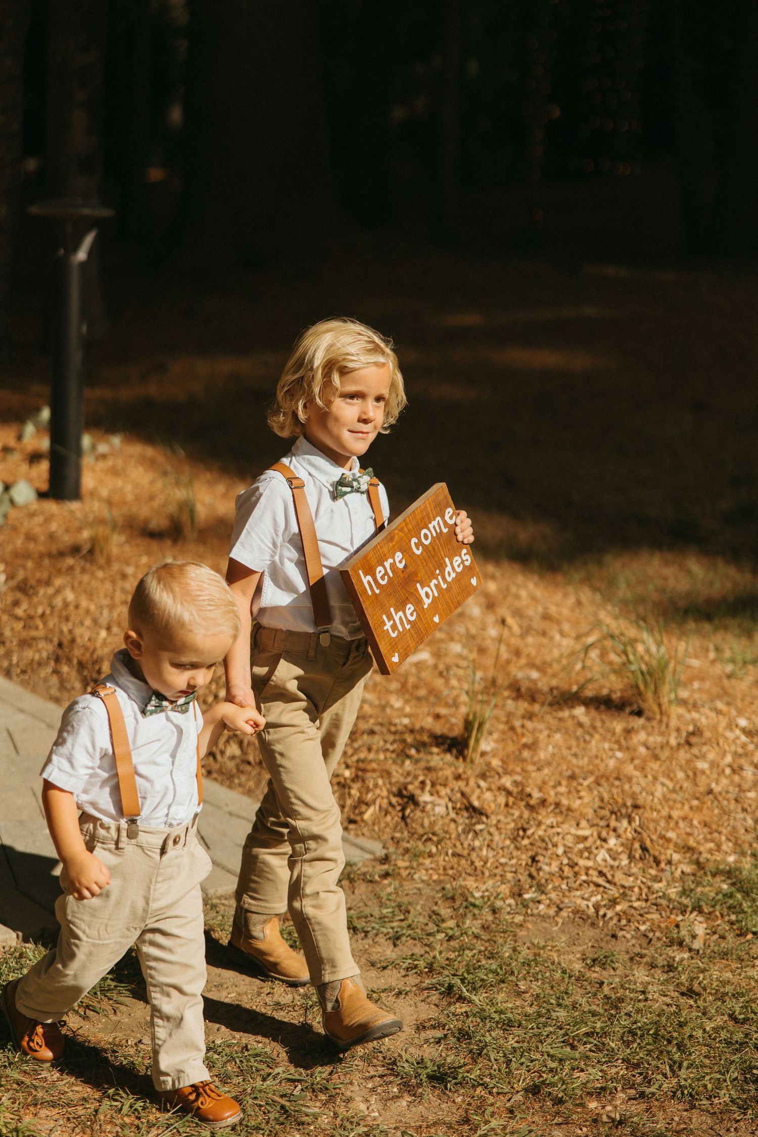 Two young ring bearers in suspenders carry a here come the brides sign at an outdoor wedding.