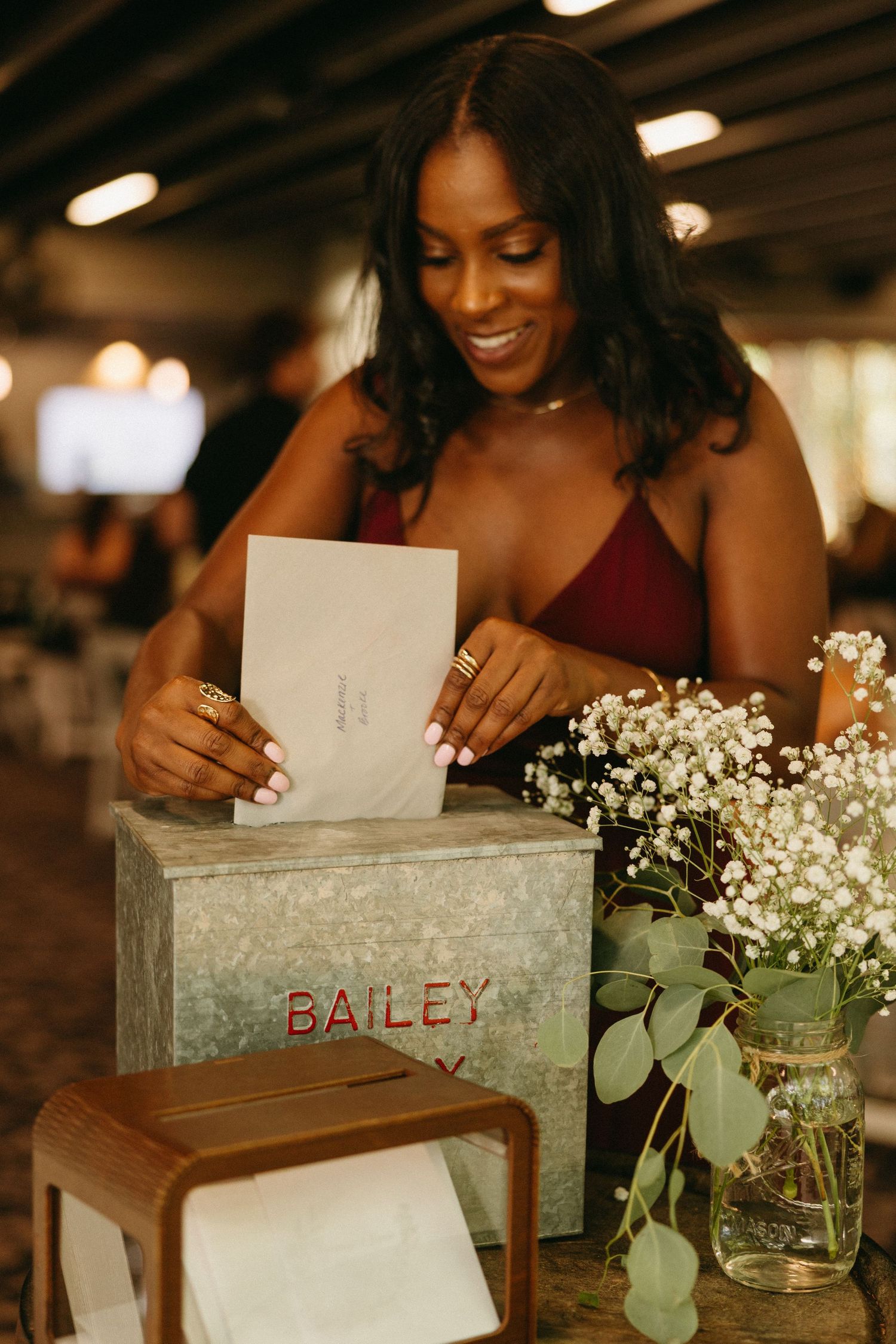 Smiling woman in red dress placing card in metal Bailey wedding card box with floral decor.
