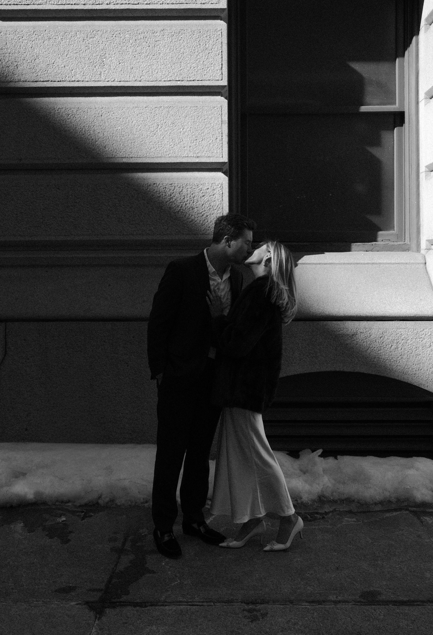 Black and white photo of a couple sharing a romantic kiss on a snowy city sidewalk at night.film-inspired