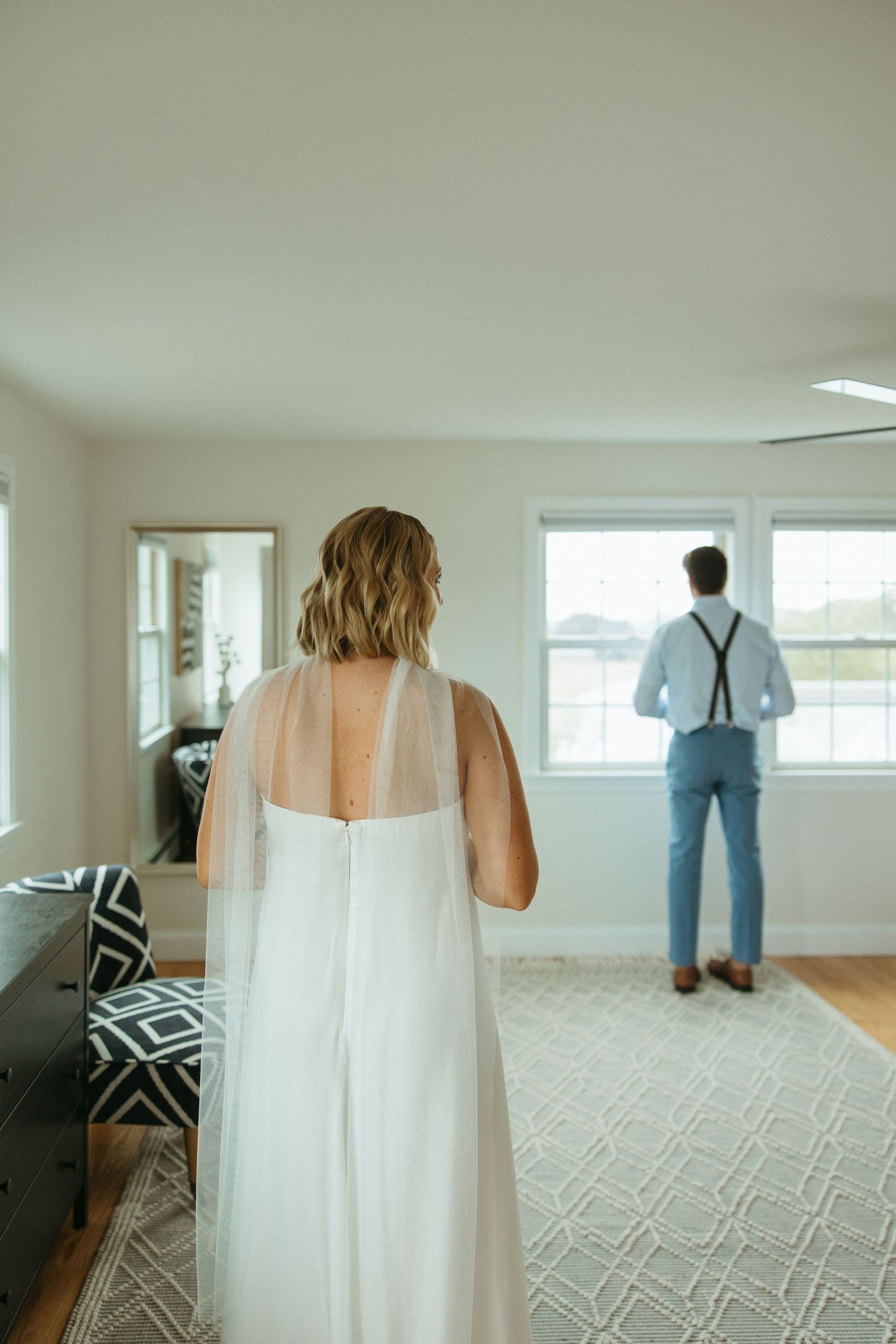 Bride in white gown with veil approaches groom in blue pants and suspenders for first look indoors.