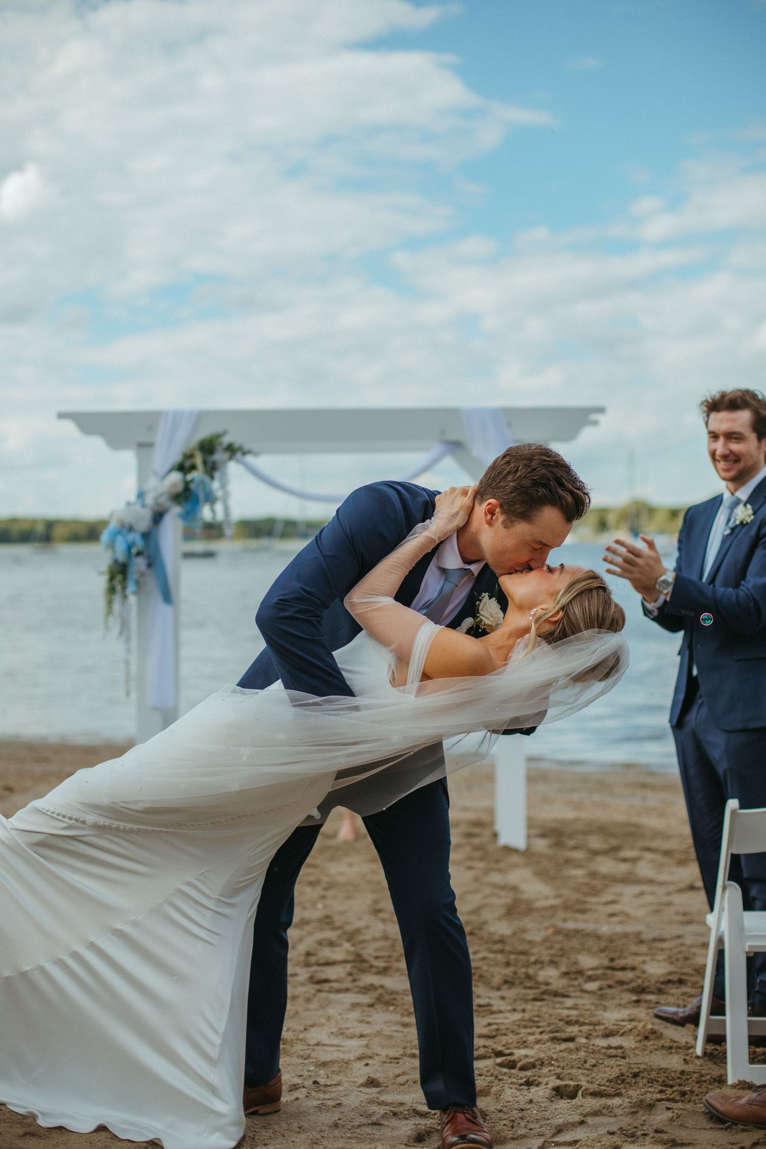 Bride and groom share a romantic dip kiss on a sandy beach during their outdoor waterfront wedding ceremony.