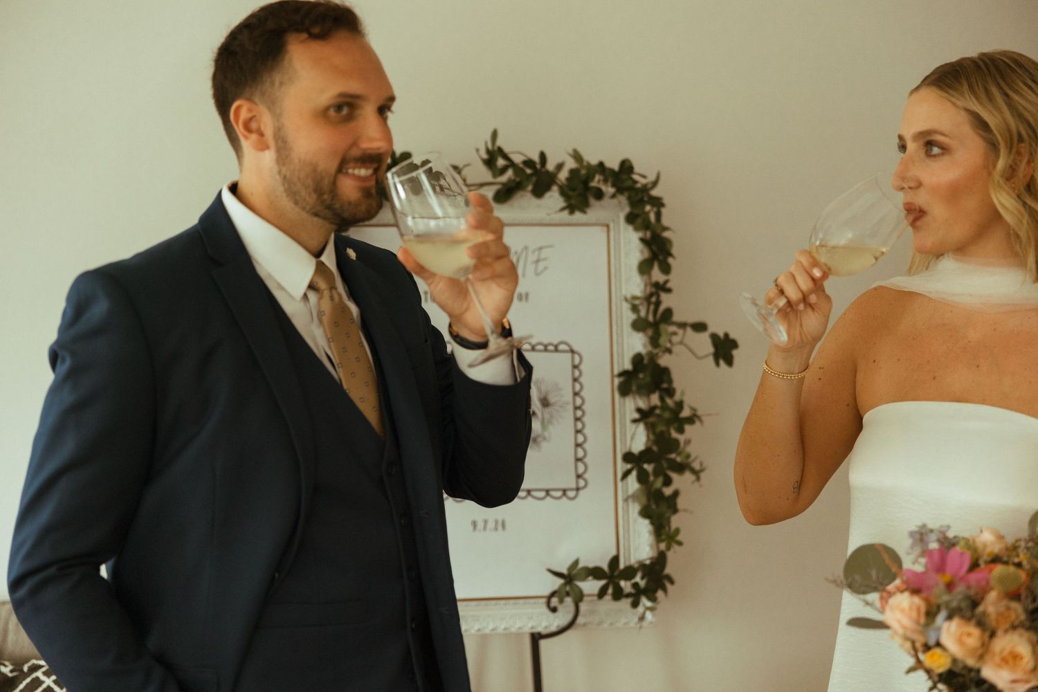 Newlyweds toast with champagne glasses indoors, groom in navy suit, bride in white wedding dress.