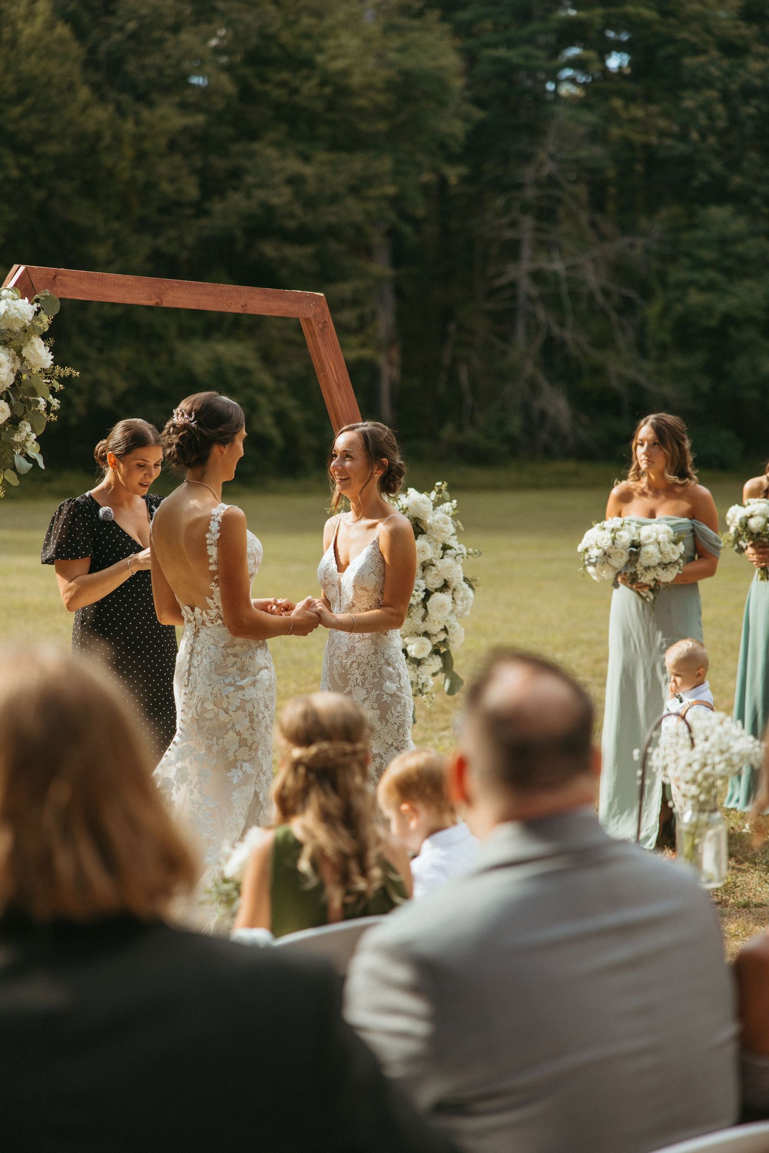Two brides exchange vows at outdoor wedding altar with wooden arch, white florals, and mint bridesmaids.