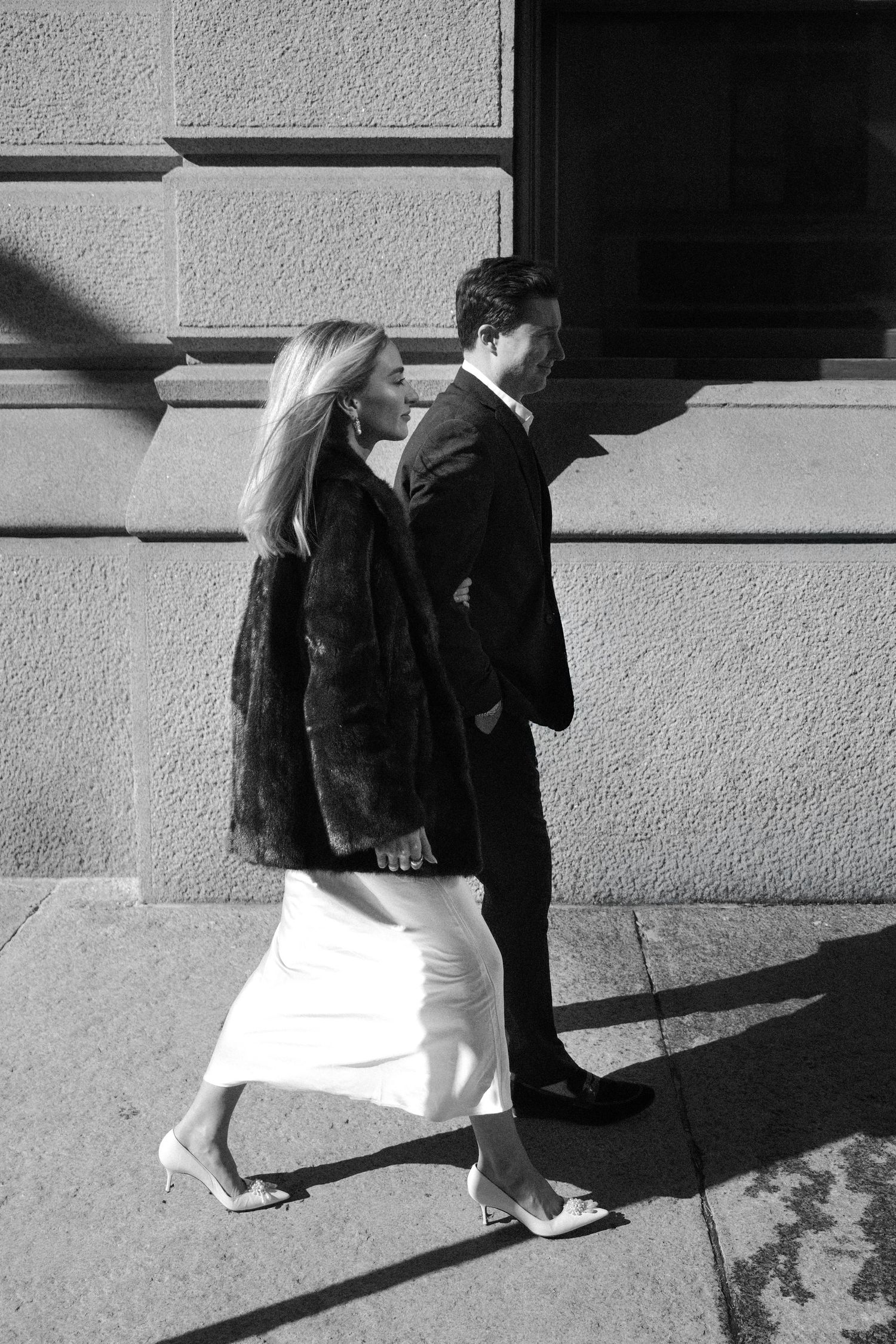 Black and white photo of a stylish couple walking past a stone building wall in bright sunlight.film-inspired