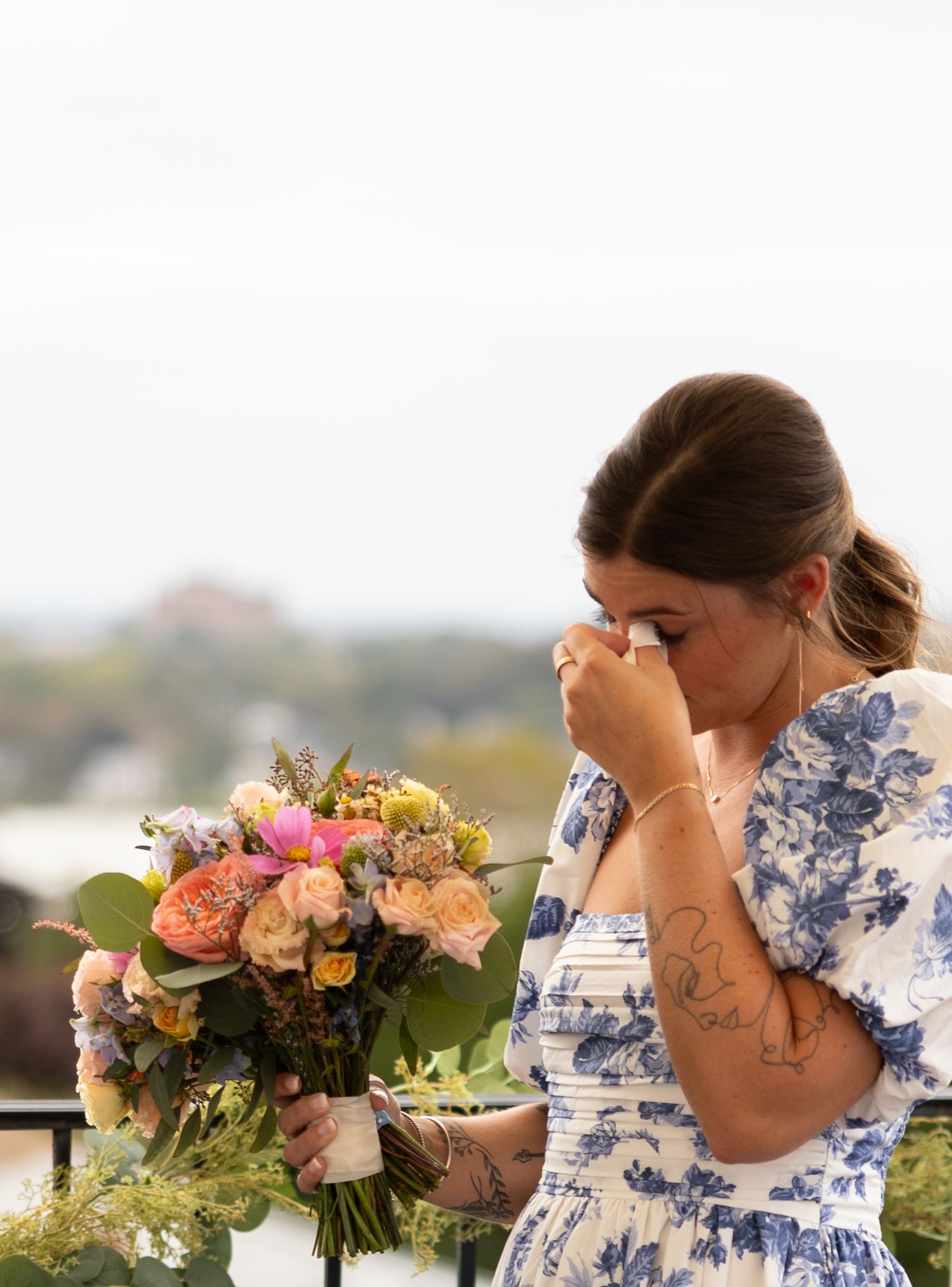 Emotional bridesmaid in floral dress wipes tears while holding a pastel wedding bouquet outdoors.