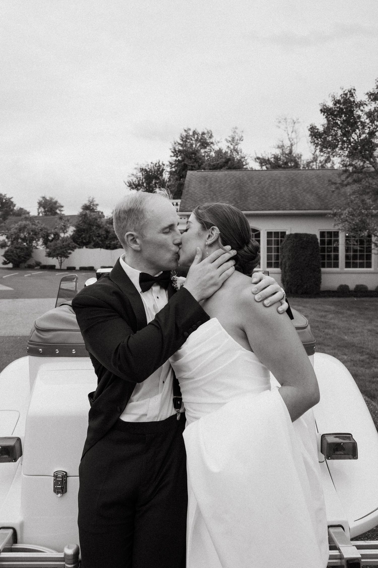 Black and white photo of bride and groom sharing a passionate kiss in front of a vintage white car.