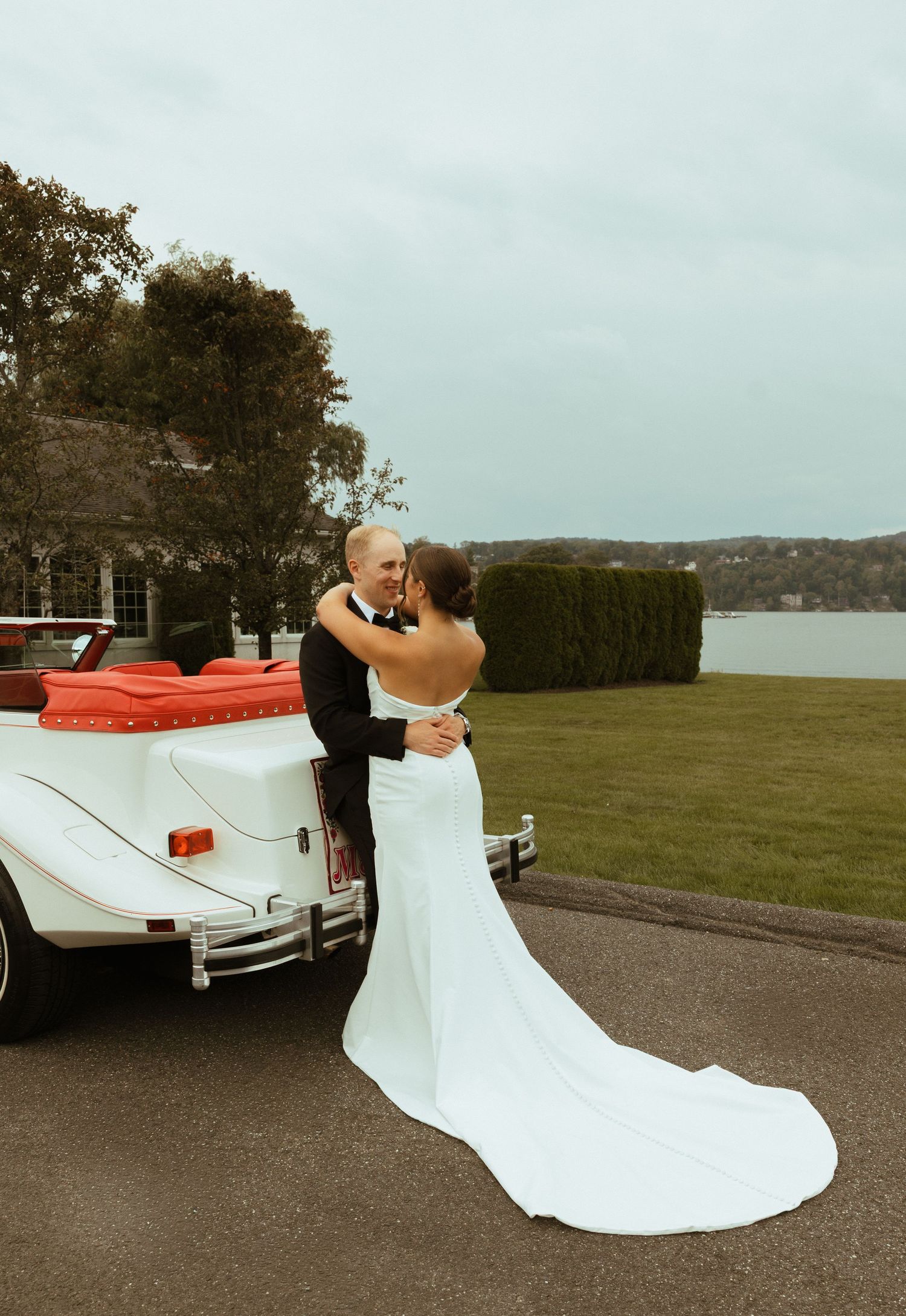 Bride and groom embracing beside a classic white convertible car with a beautiful lakeside backdrop.