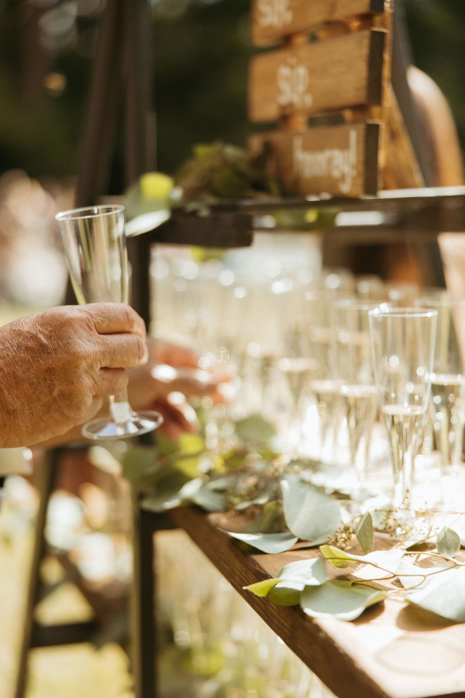 A hand reaches for a champagne flute at an outdoor wedding bar decorated with eucalyptus leaves.