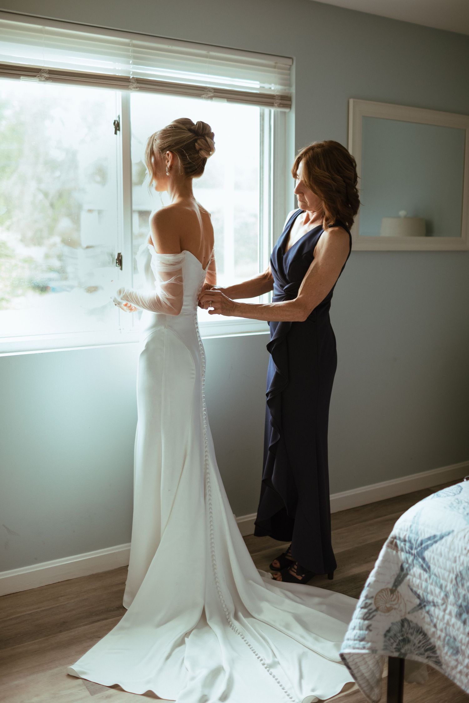 Mother helping bride button up elegant off-shoulder white wedding gown with train by window.