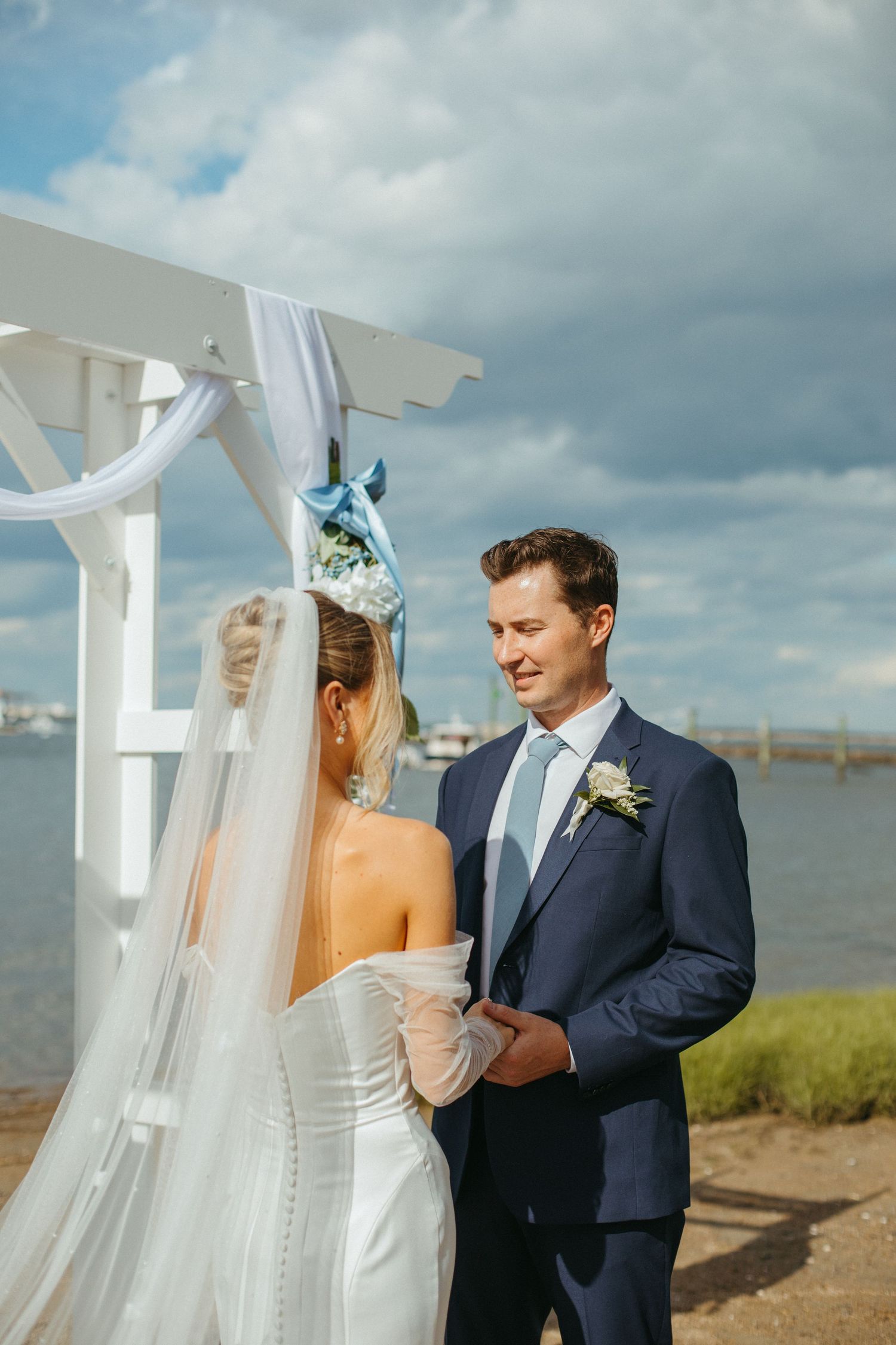 Bride and groom holding hands at white waterside wedding altar with blue ribbon decorations and cloudy sky.