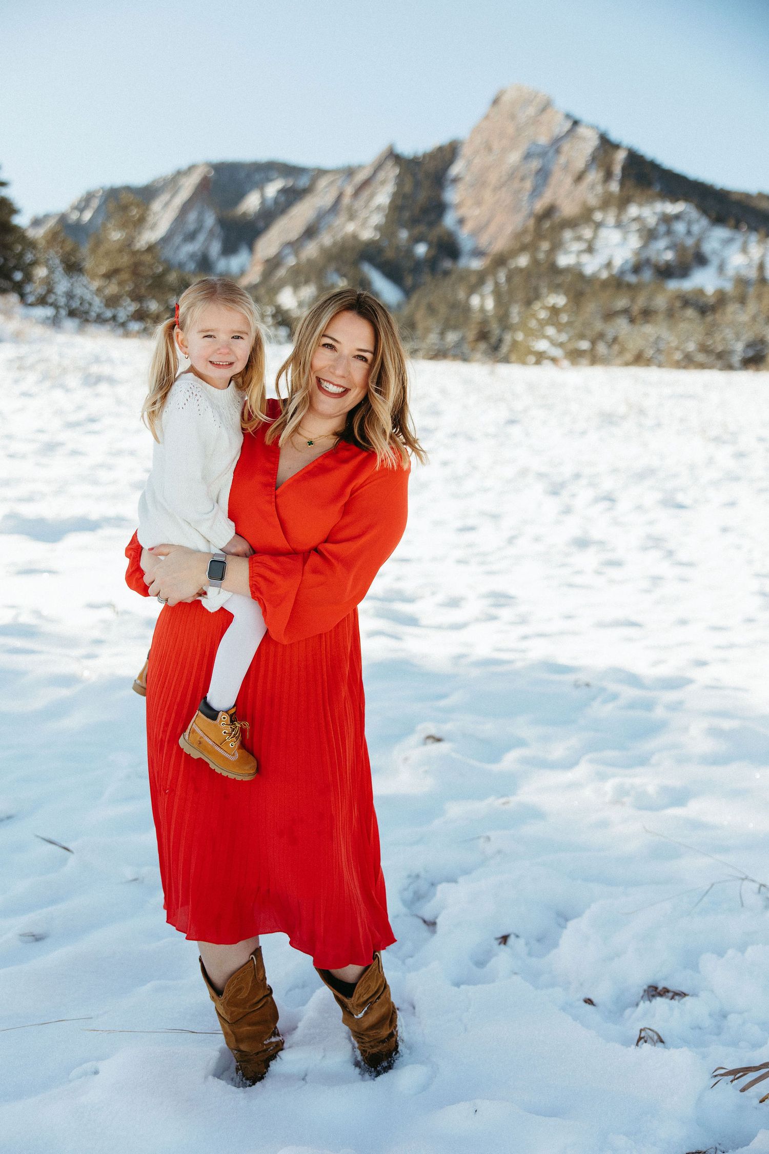 Woman in red dress holds young girl in snowy Colorado mountain landscape with rocky flatirons in background.