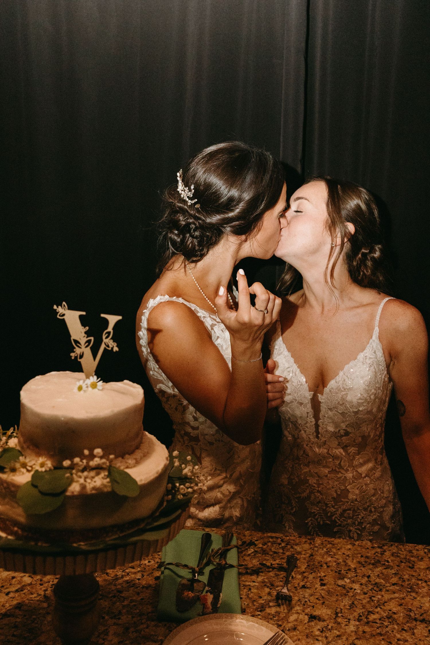 Two brides share a kiss beside their two-tiered white wedding cake.film-inspired