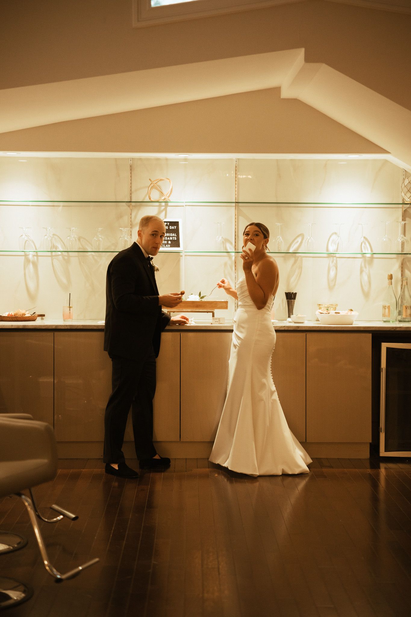 Bride in white gown and groom in tuxedo at a wedding reception bar.