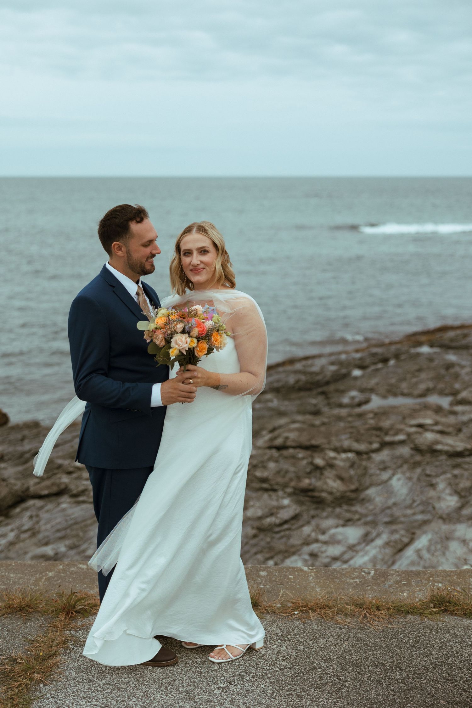 Bride and groom embrace by the rocky ocean shore, she holds a colorful bouquet while wearing a flowing white gown.