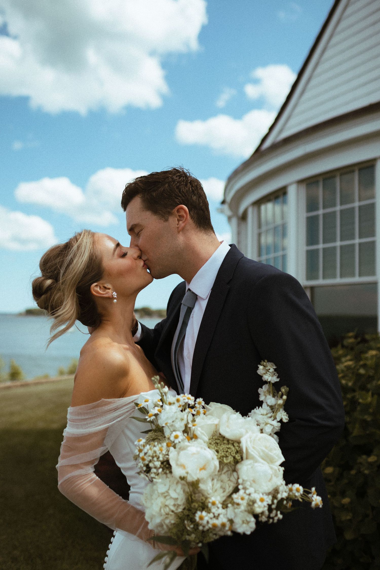 Bride and groom sharing kiss outdoors holding white floral bouquet by waterfront.