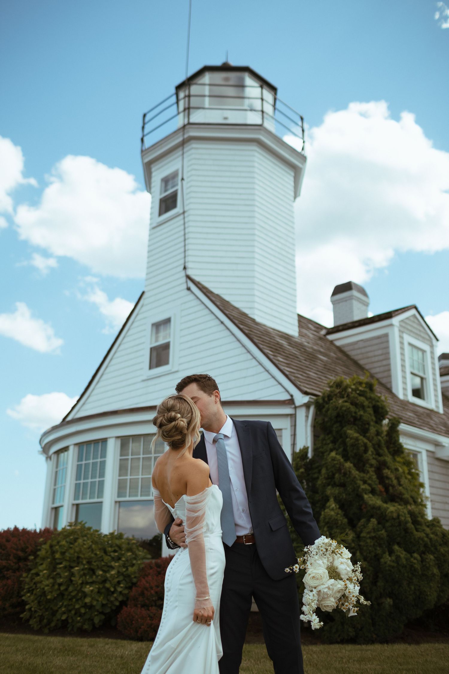 Bride and groom gaze at each other in front of a tall white lighthouse on a beautiful sunny wedding day.