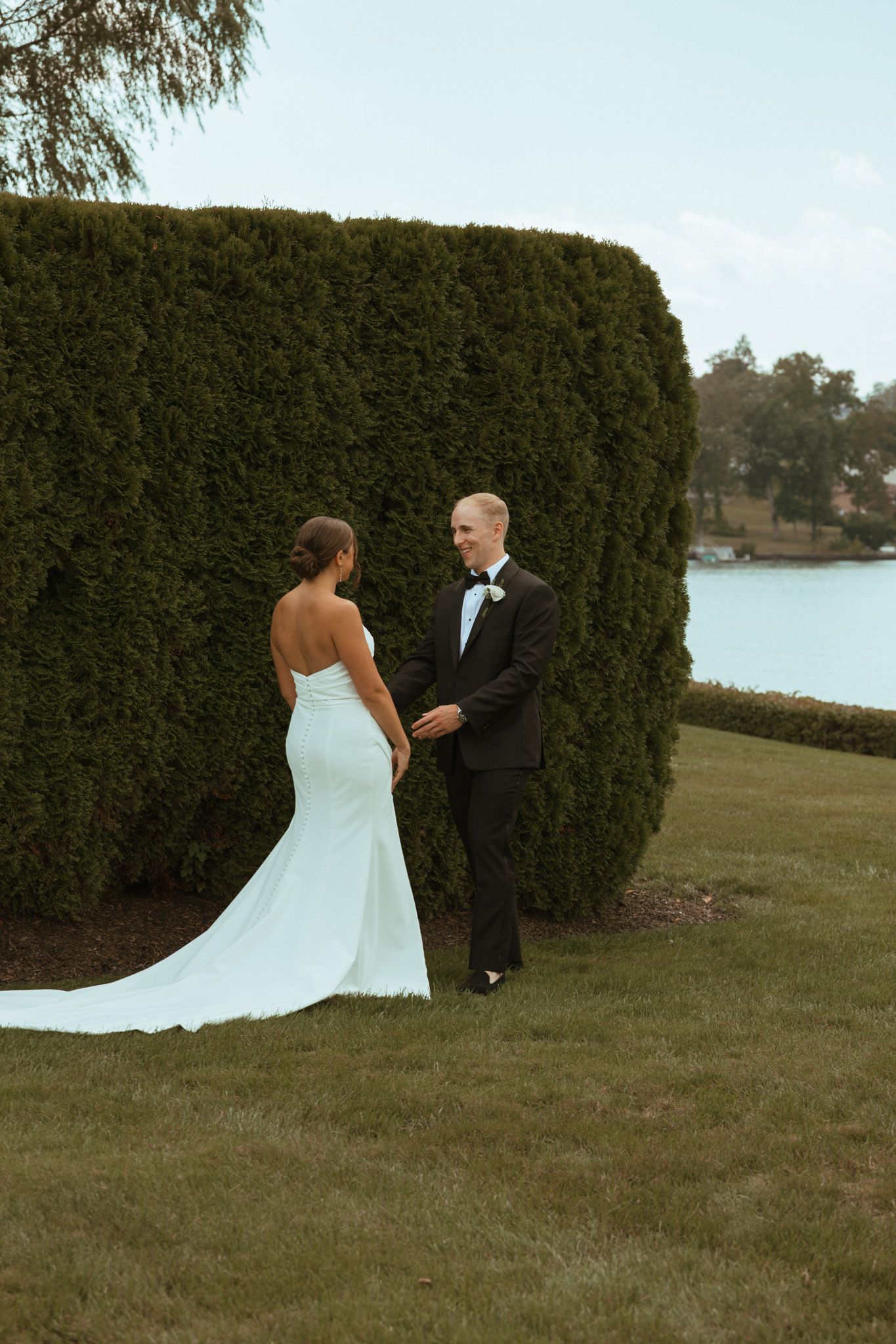 Bride and groom sharing first look moment beside tall green hedge near waterfront.