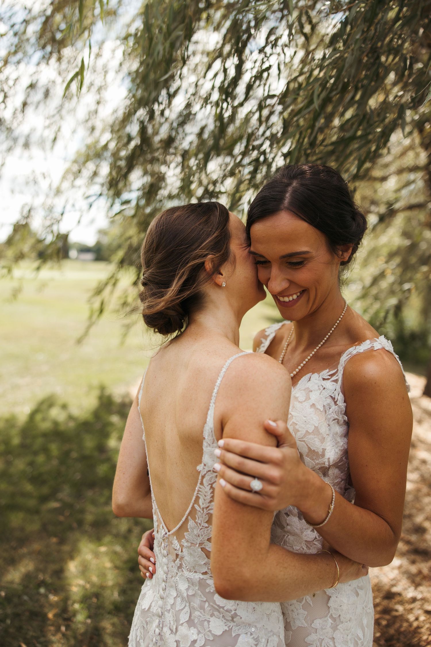 Two brides in lace wedding dresses share a joyful embrace under weeping willow branches outdoors.