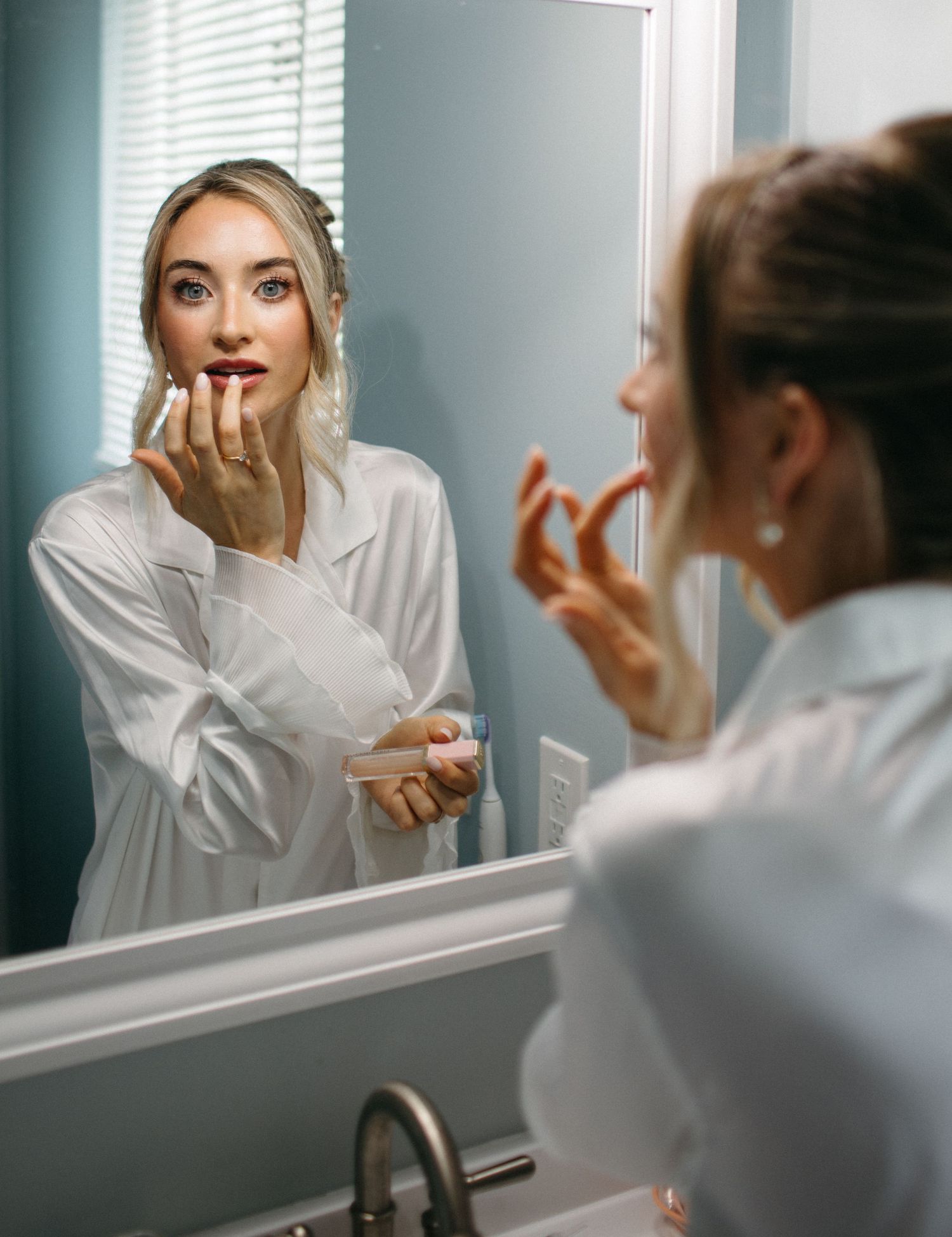 Bride applying red lipstick while looking in bathroom mirror wearing white satin robe on wedding day.