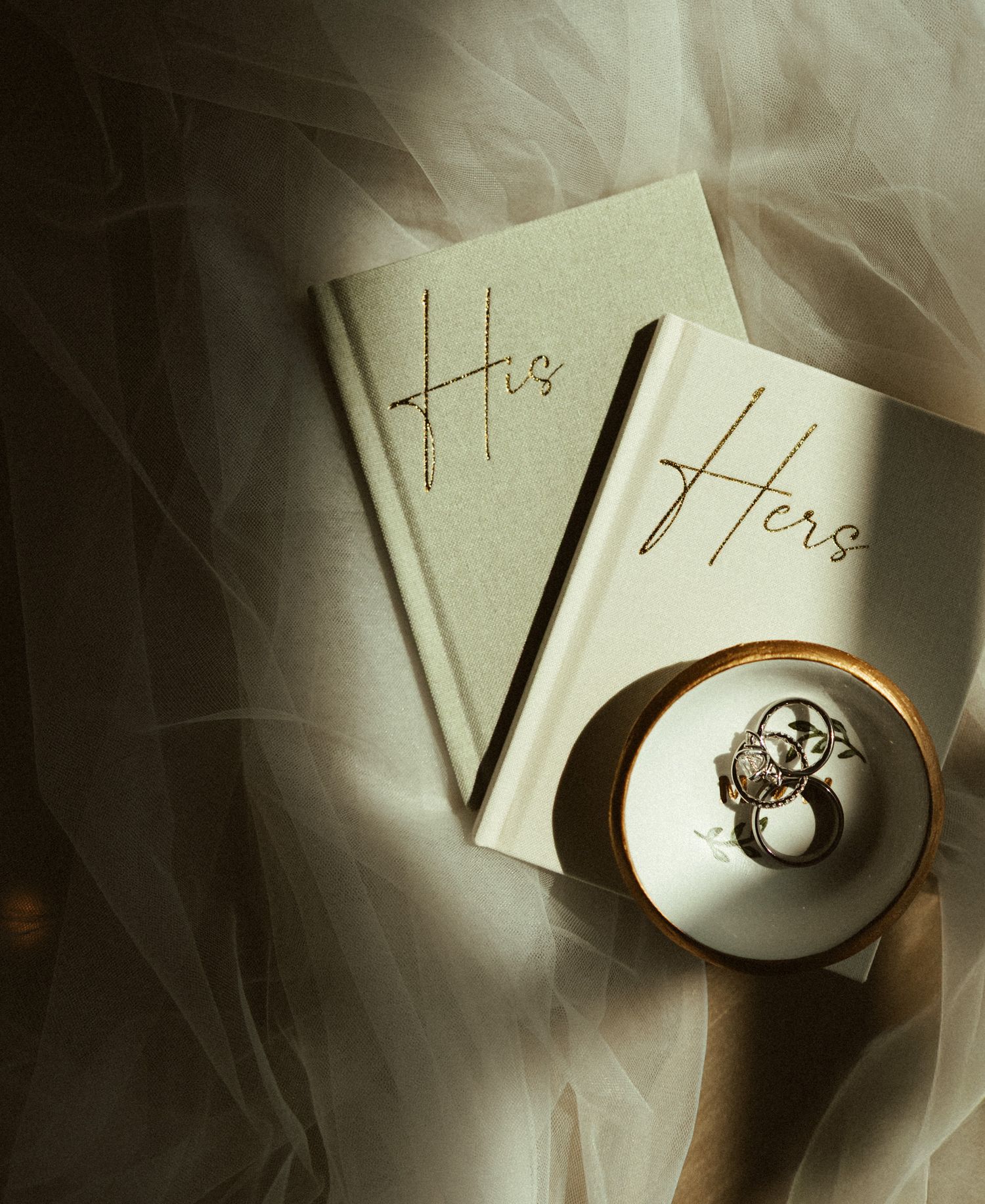 His and Hers wedding vow books with rings on a decorative dish against sheer fabric.