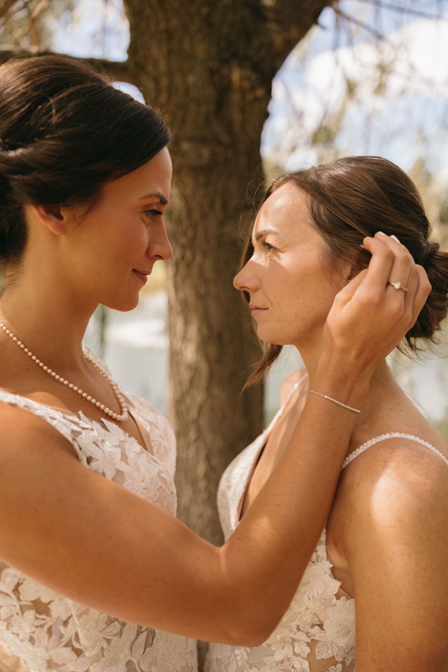 Two brides in lace wedding gowns share an intimate moment, one tenderly touching the others hair outdoors.