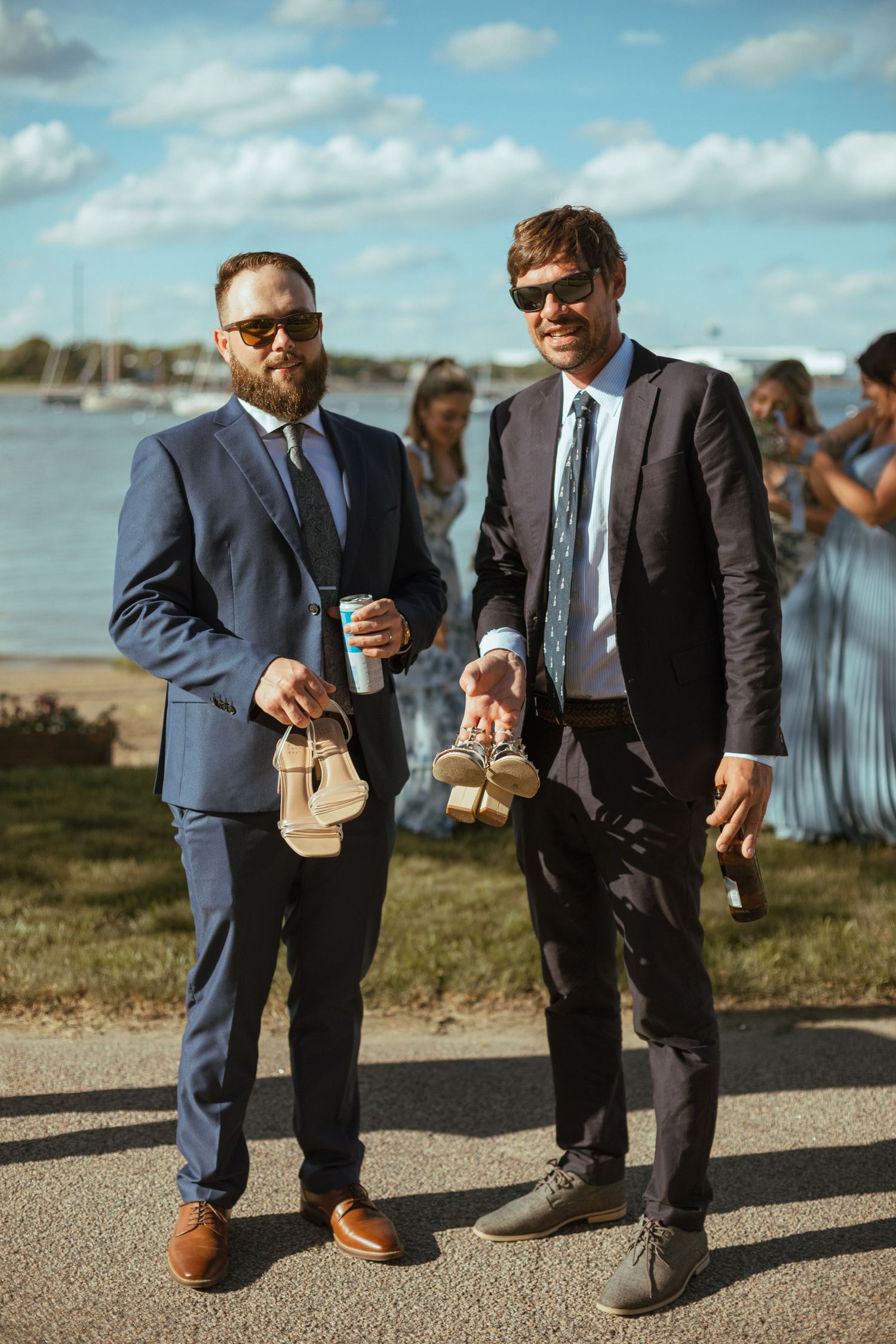 Two groomsmen in navy suits holding shoes and drinks outdoors at waterfront wedding reception.