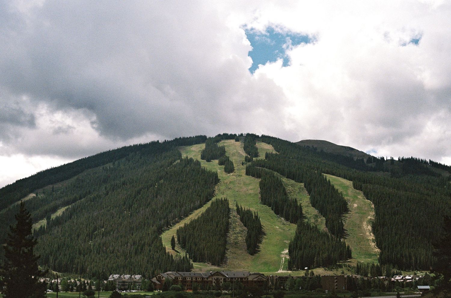 Scenic summer mountain landscape with ski runs visible on green forested slopes under dramatic cloudy skies.
