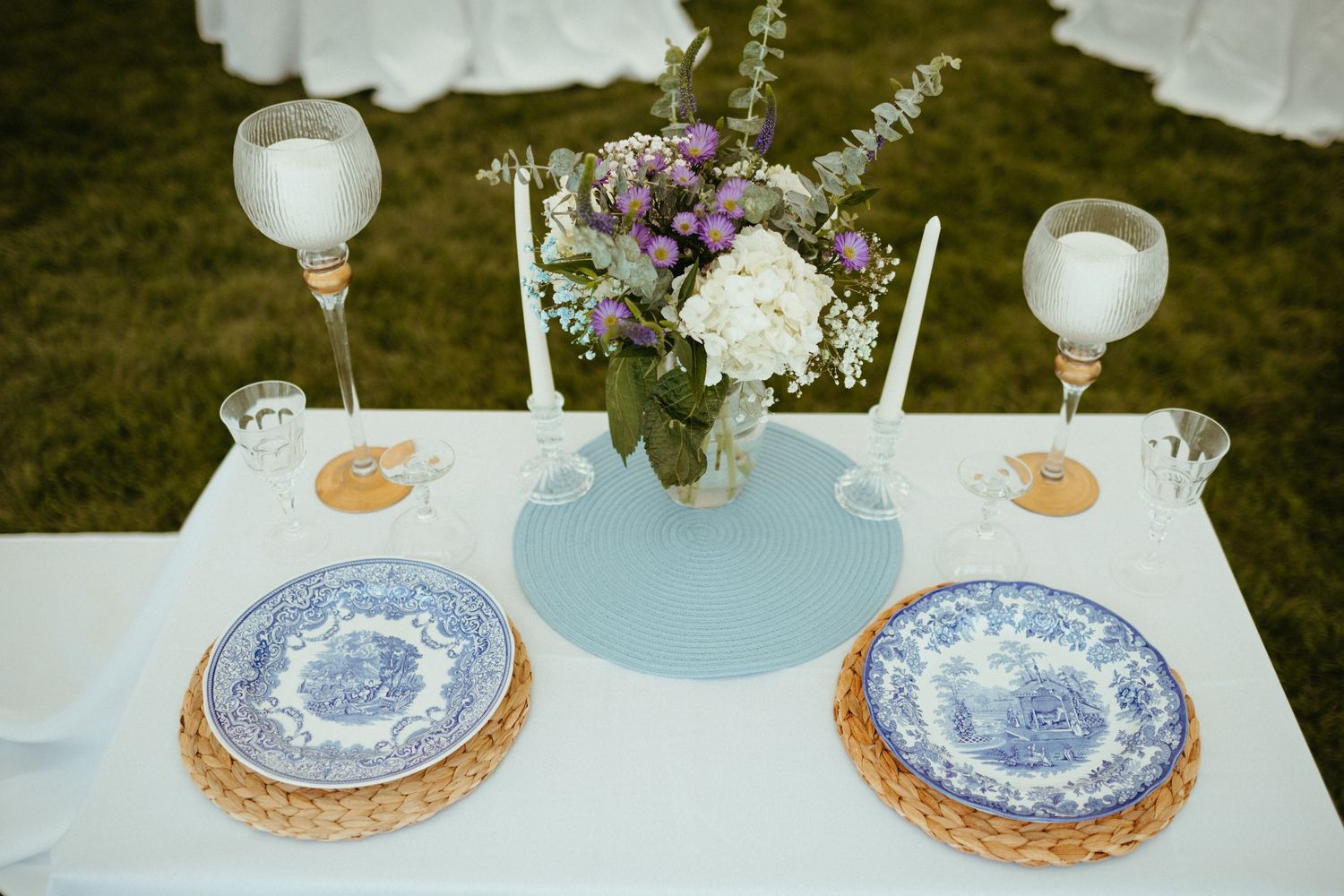 Elegant wedding table with blue transferware plates, wicker chargers, candles, and purple floral centerpiece.