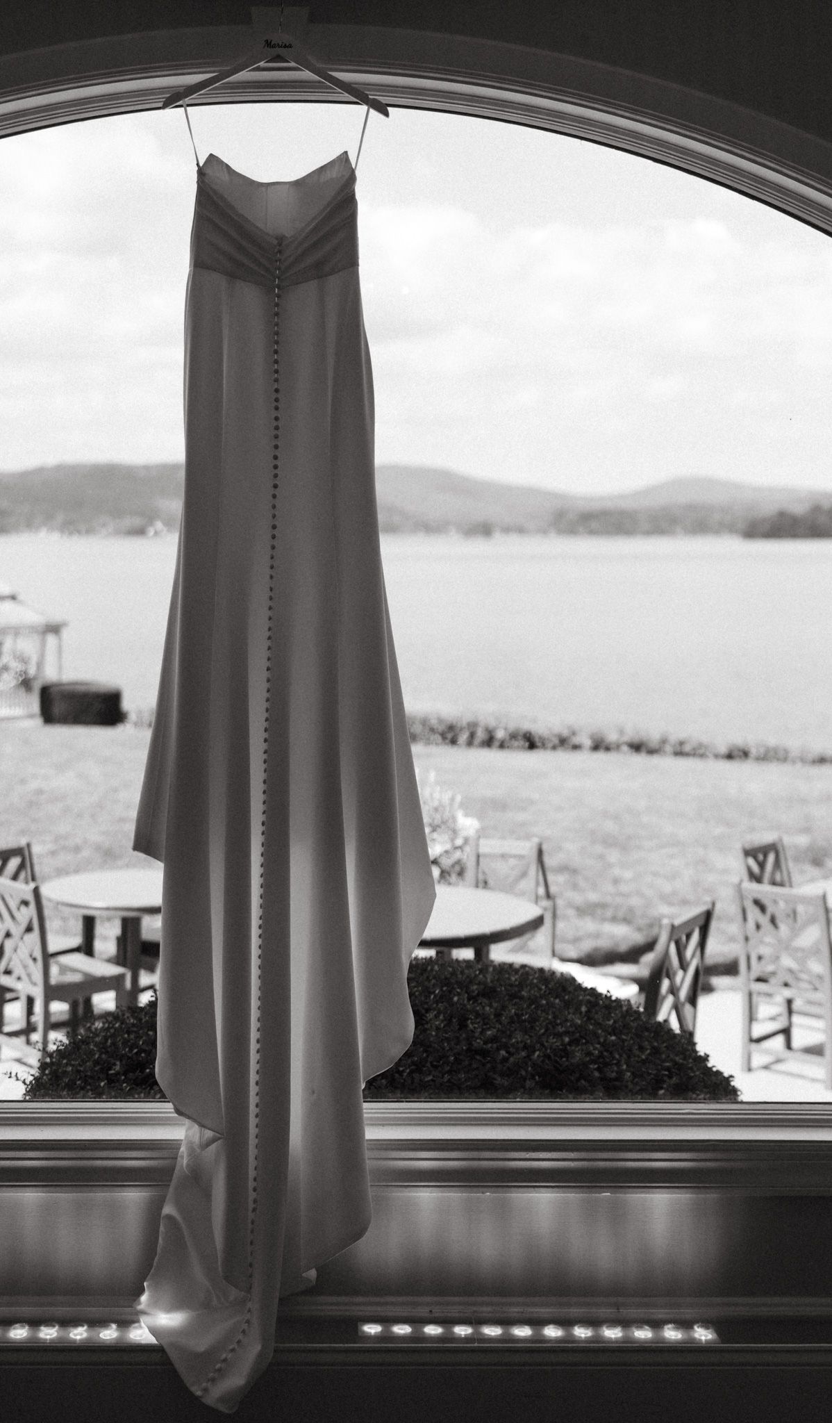 Black and white photo of elegant wedding gown hanging in arched window with lake view.