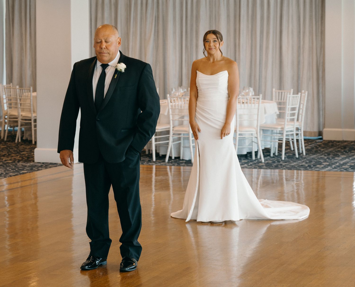 Bride in fitted white gown standing with father in black suit on dance floor.
