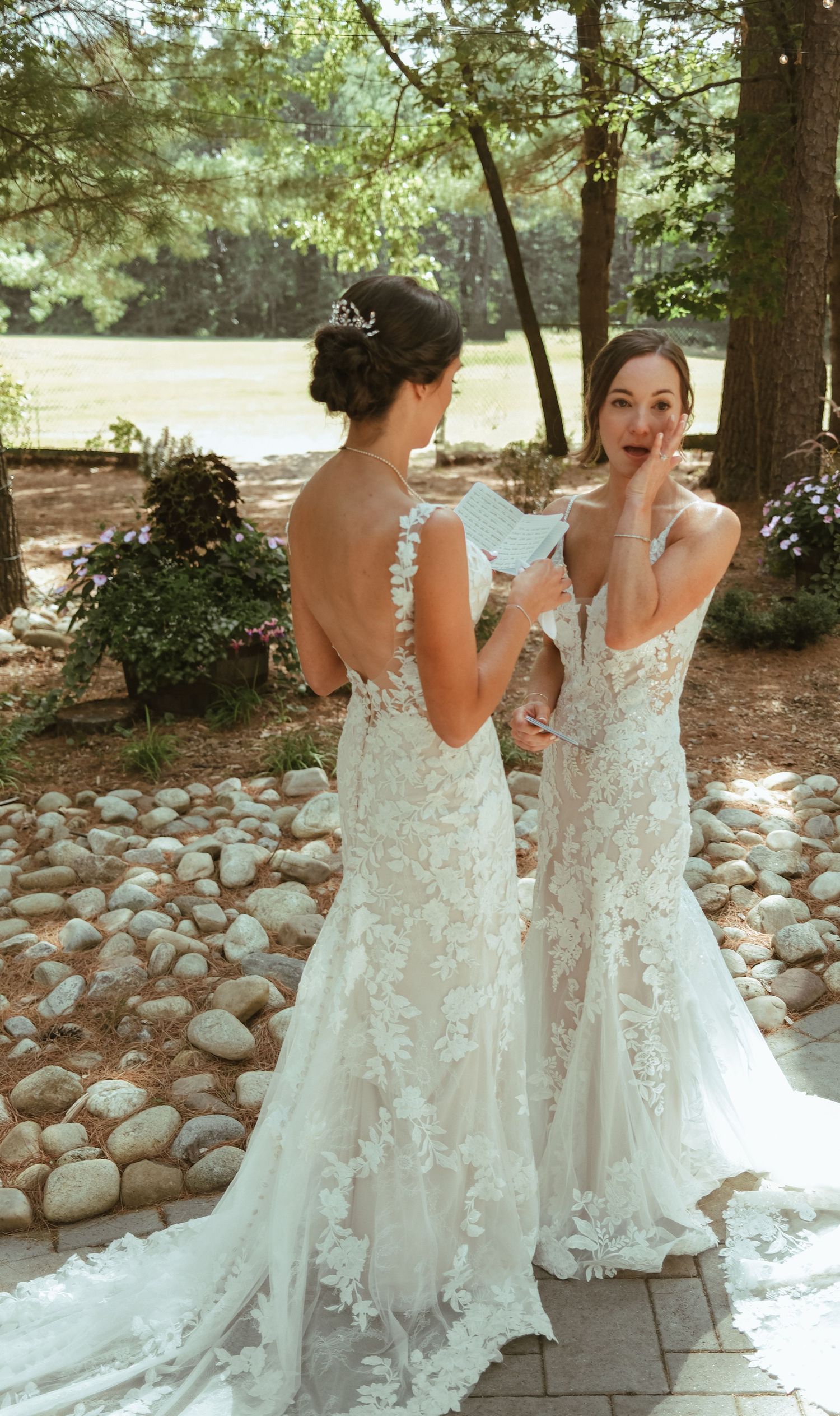 Two brides in lace wedding gowns exchange vows in a garden ceremony surrounded by trees and stones.