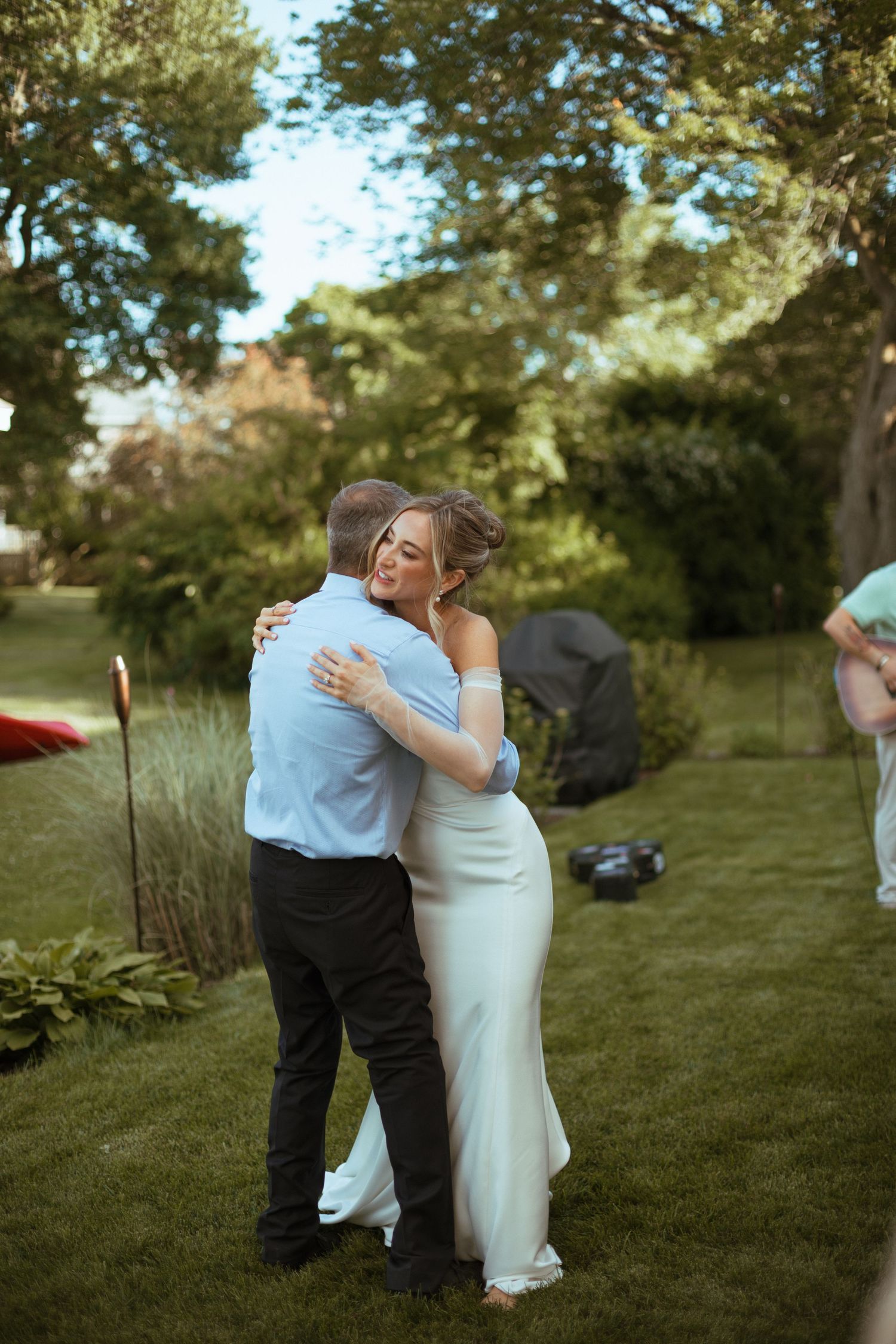 Bride in white gown sharing tender embrace with guest on green lawn at outdoor wedding.