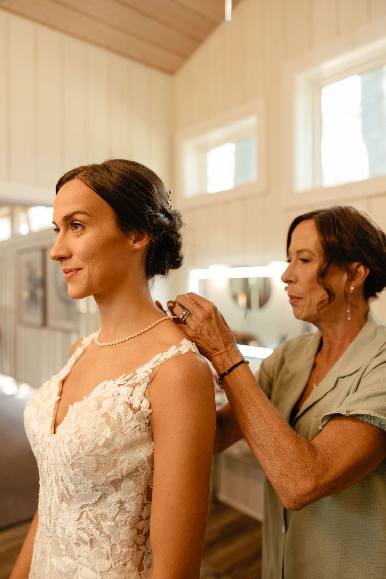 Older woman in sage green dress fastens pearl necklace on bride in lace wedding gown before ceremony.