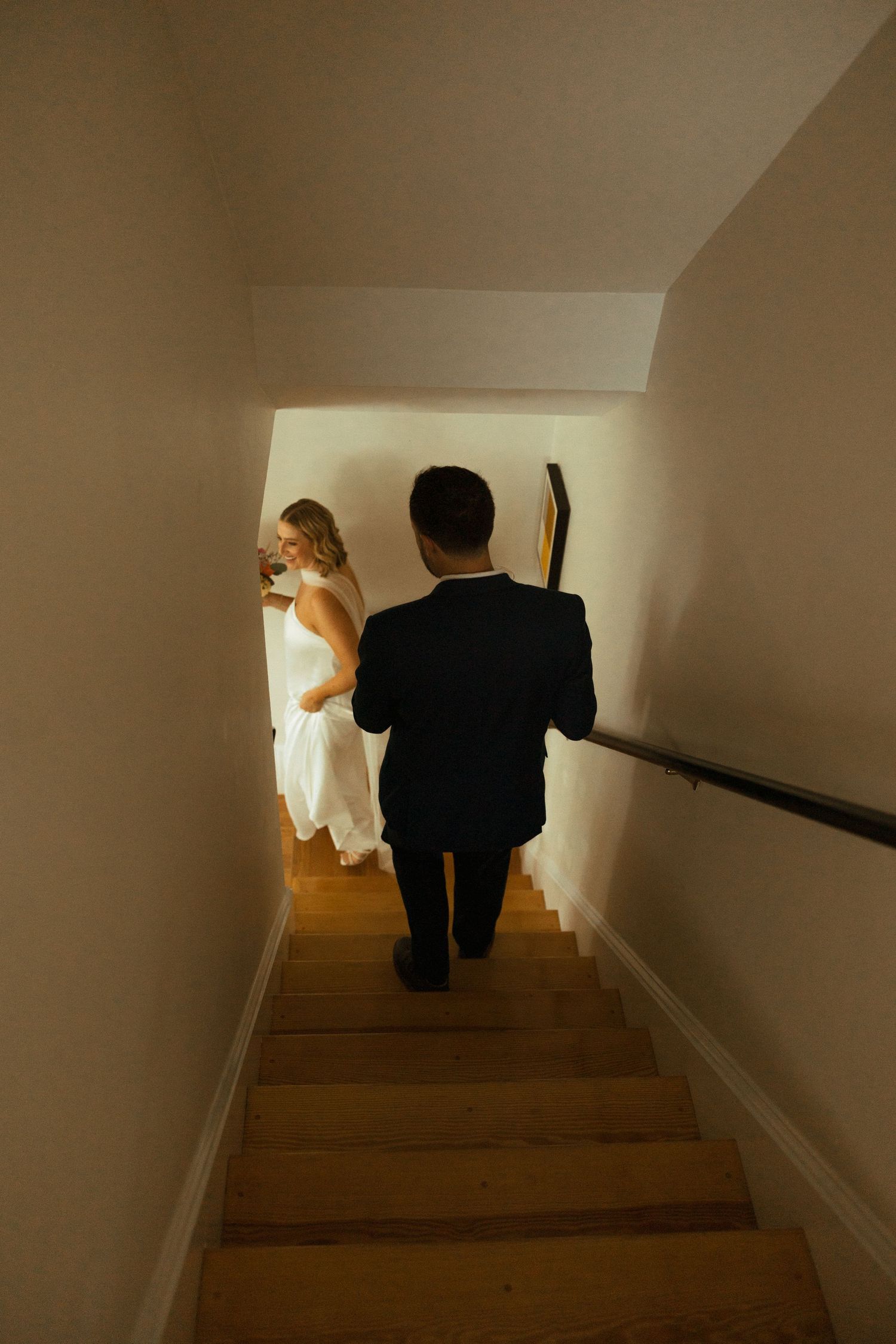 Bride and groom walking up wooden staircase together in wedding attire from behind.