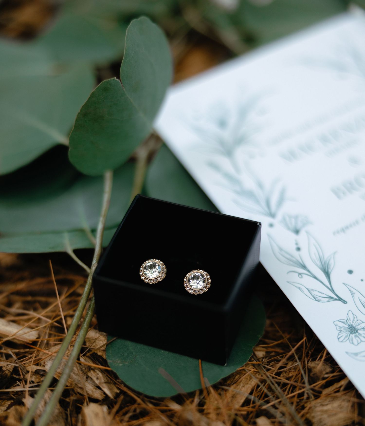 Round diamond halo stud earrings in black jewelry box beside eucalyptus leaves and wedding invitation.