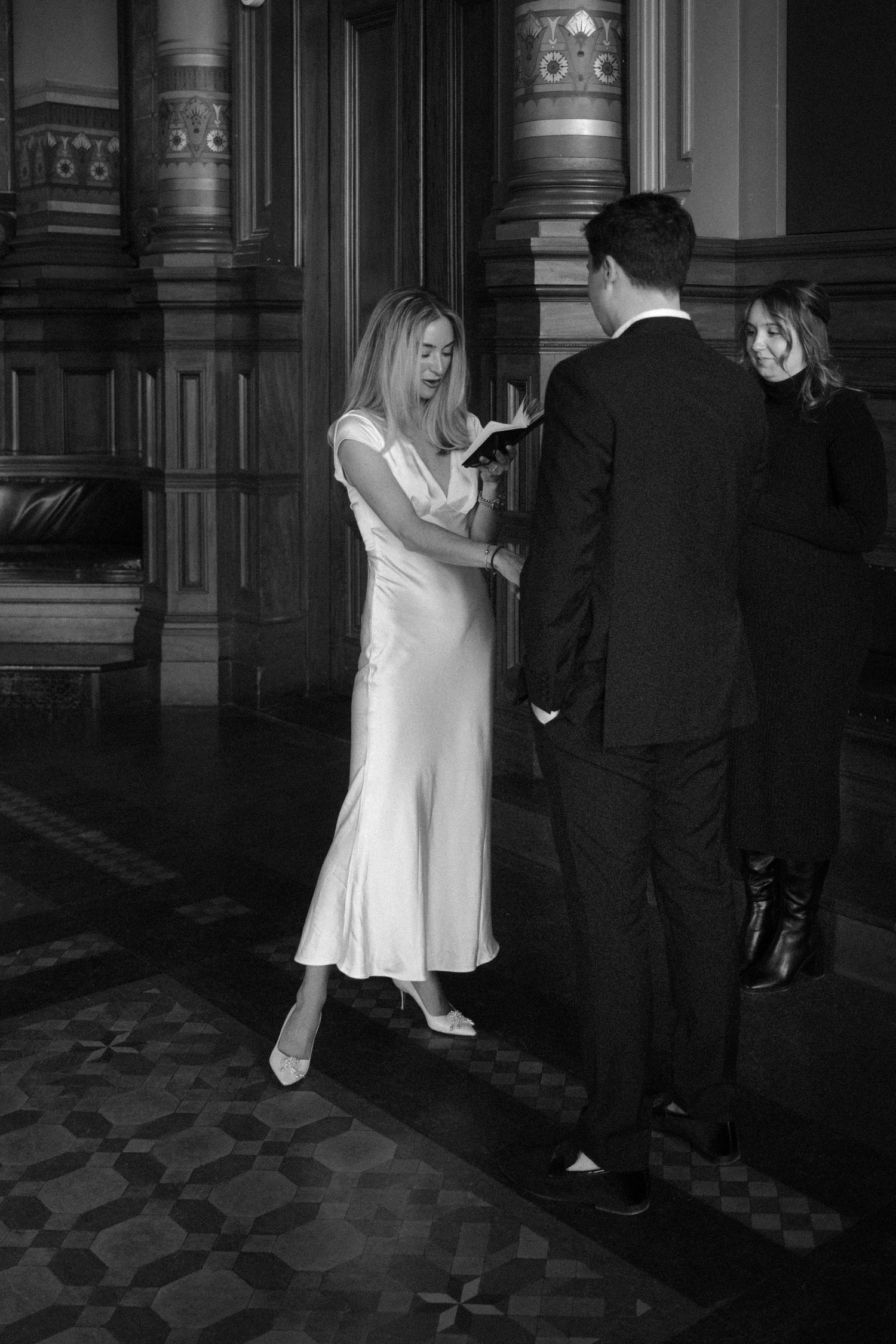 Bride in white satin dress exchanging rings with groom in elegant hall.film-inspired