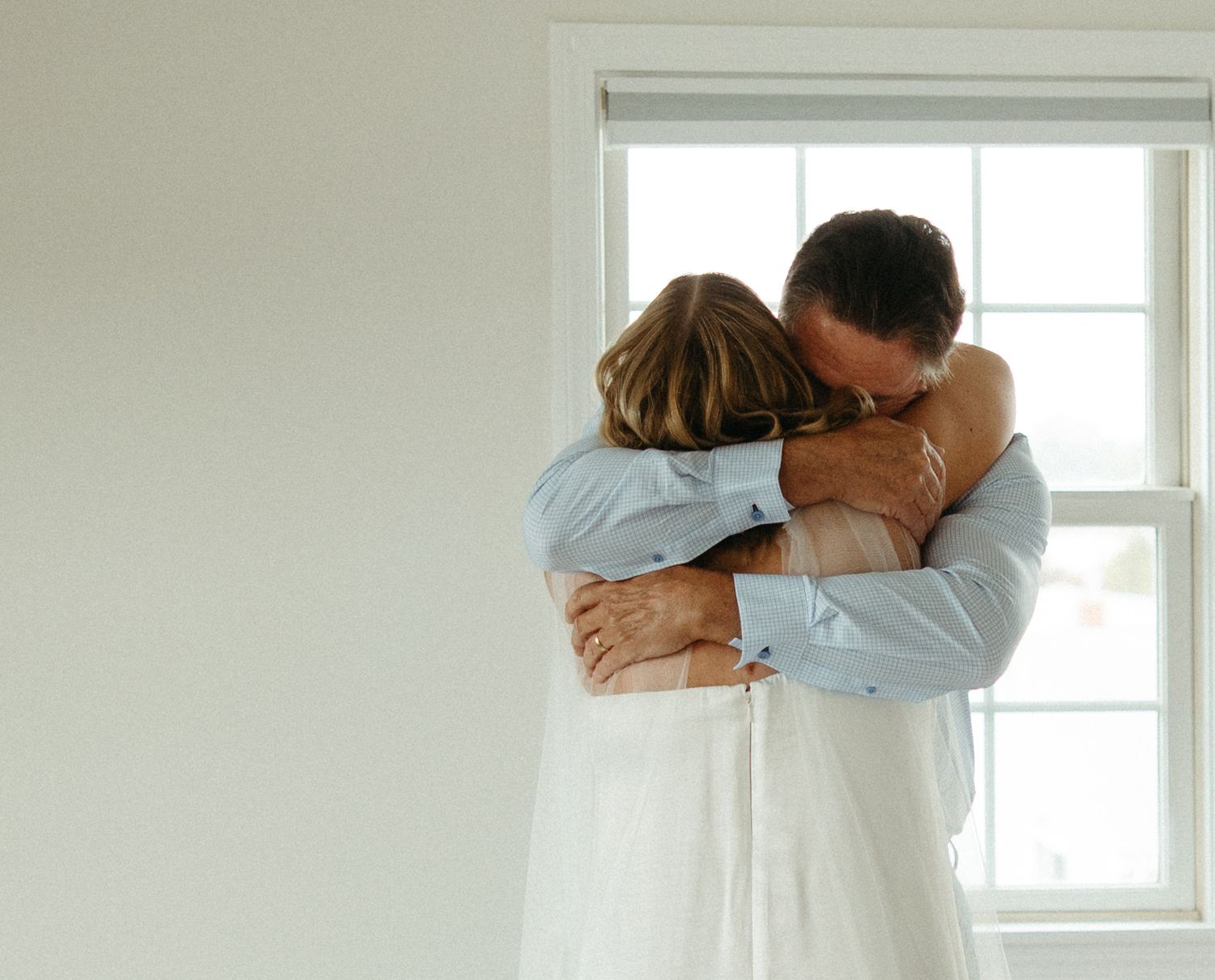 Bride and father share an emotional embrace near a bright window before the wedding ceremony.